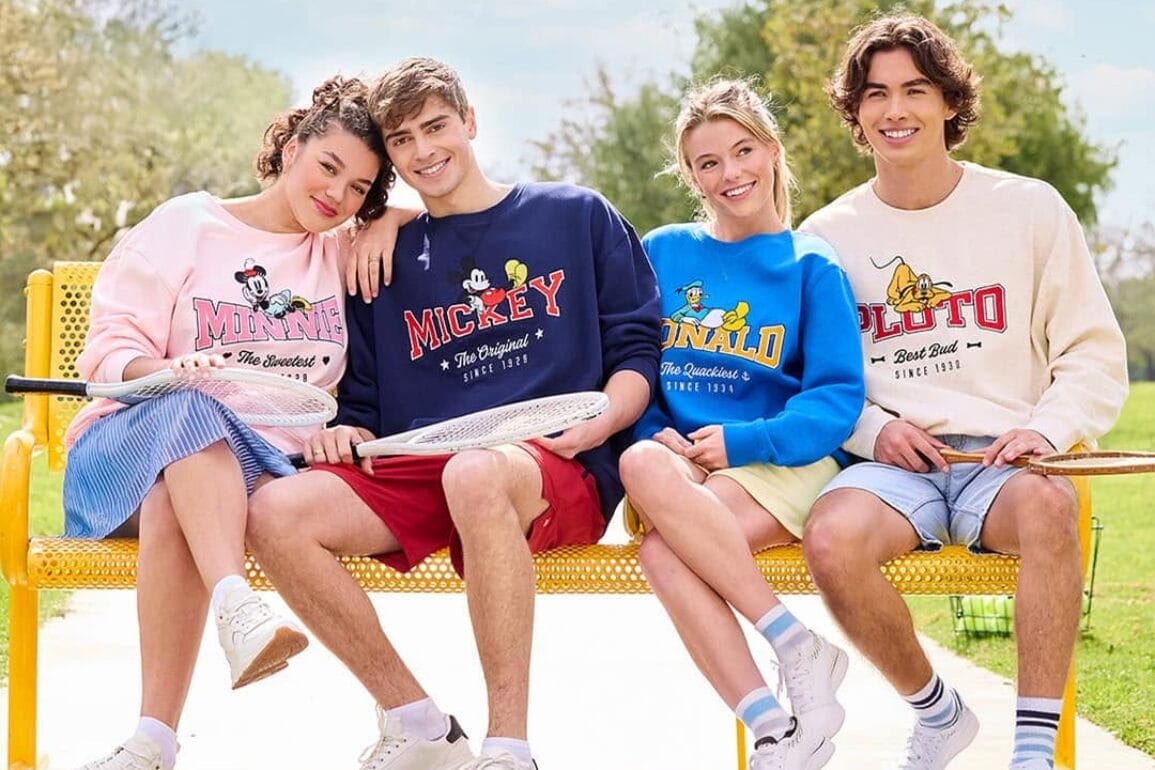 Four people sit on a bench outdoors, all wearing colorful Mickey and friends sweatshirts featuring characters like Minnie, Mickey, Donald, and Pluto, each holding a badminton racket.