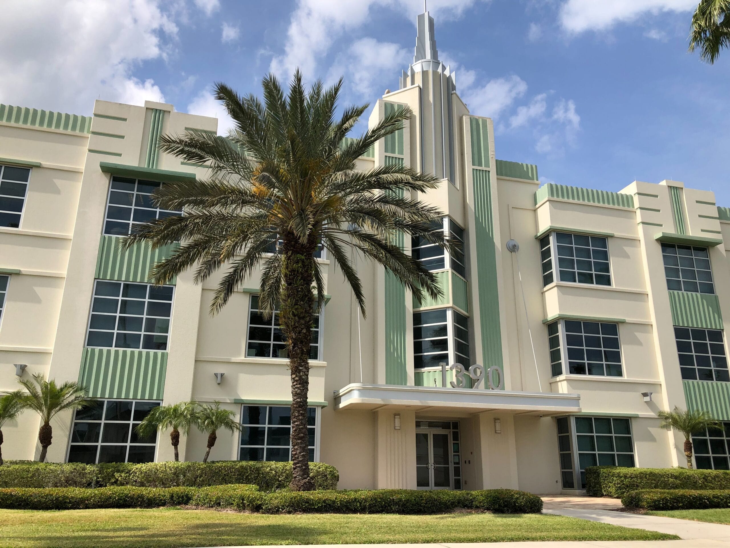 An Art Deco-style office building with green accents, large windows, and a central spire. Palm trees and well-maintained shrubs line the entrance under a clear blue sky, creating a setting fit for a celebration.