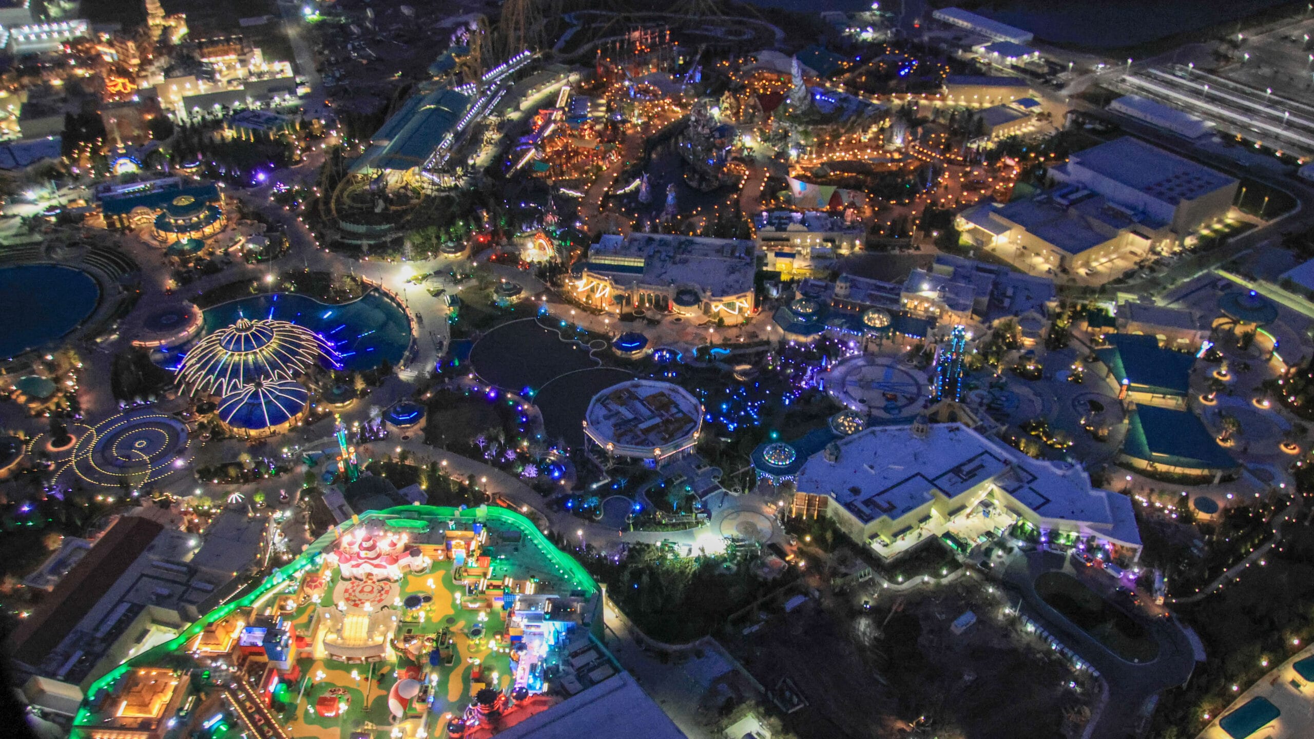 Epic Universe comes alive under dazzling night lighting, offering an aerial view of its brightly lit amusement park. Roller coasters twist and turn beside circular rides, as the park's illuminated attractions shine against a backdrop of numerous surrounding buildings.