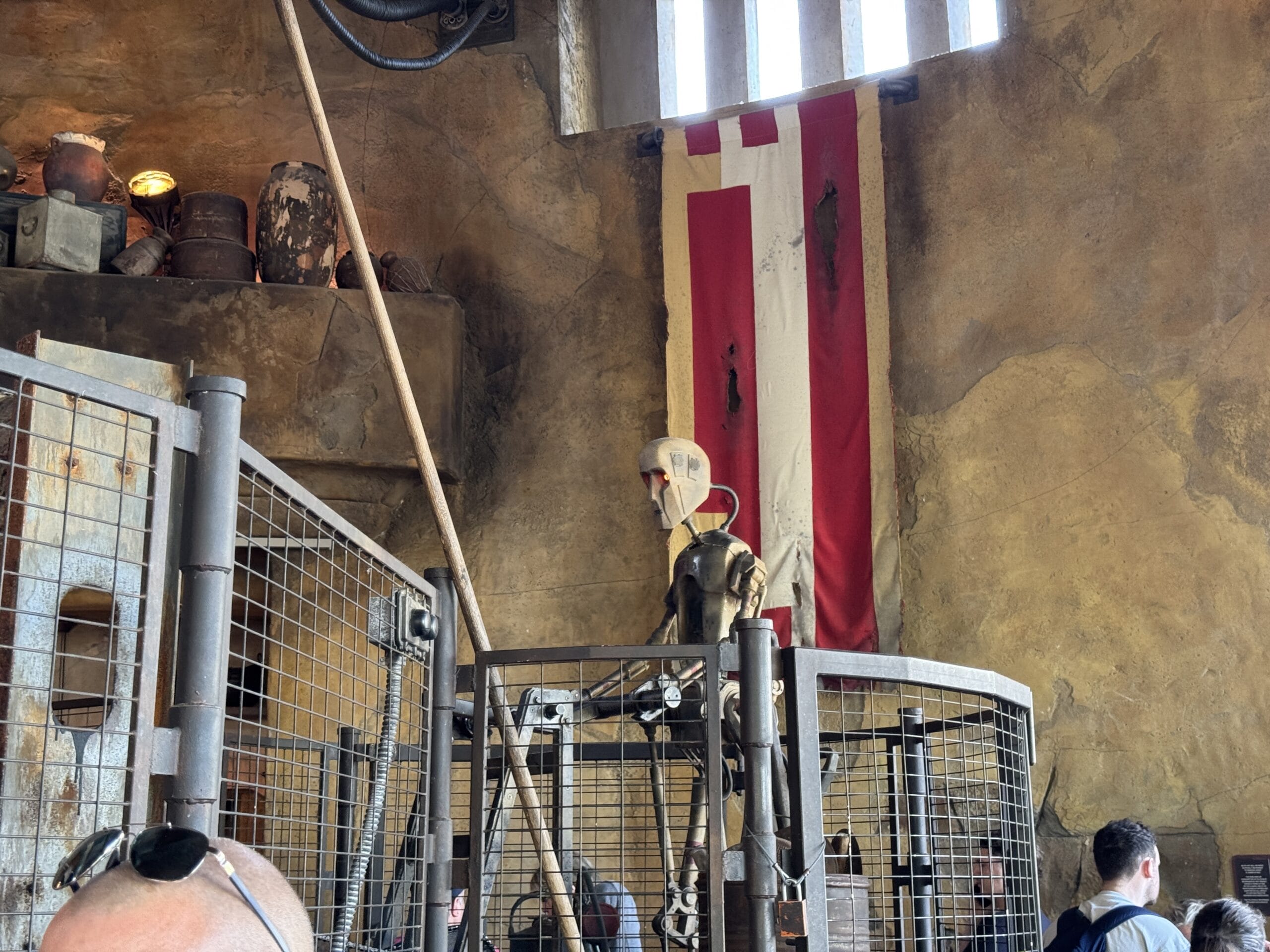 A beige humanoid robot, known as 8J-8D, stands inside a metal cage with its face fixed next to a worn flag. The scene has an industrial, rustic look straight out of Hollywood Studios, with various objects neatly arranged on shelves in the backdrop.