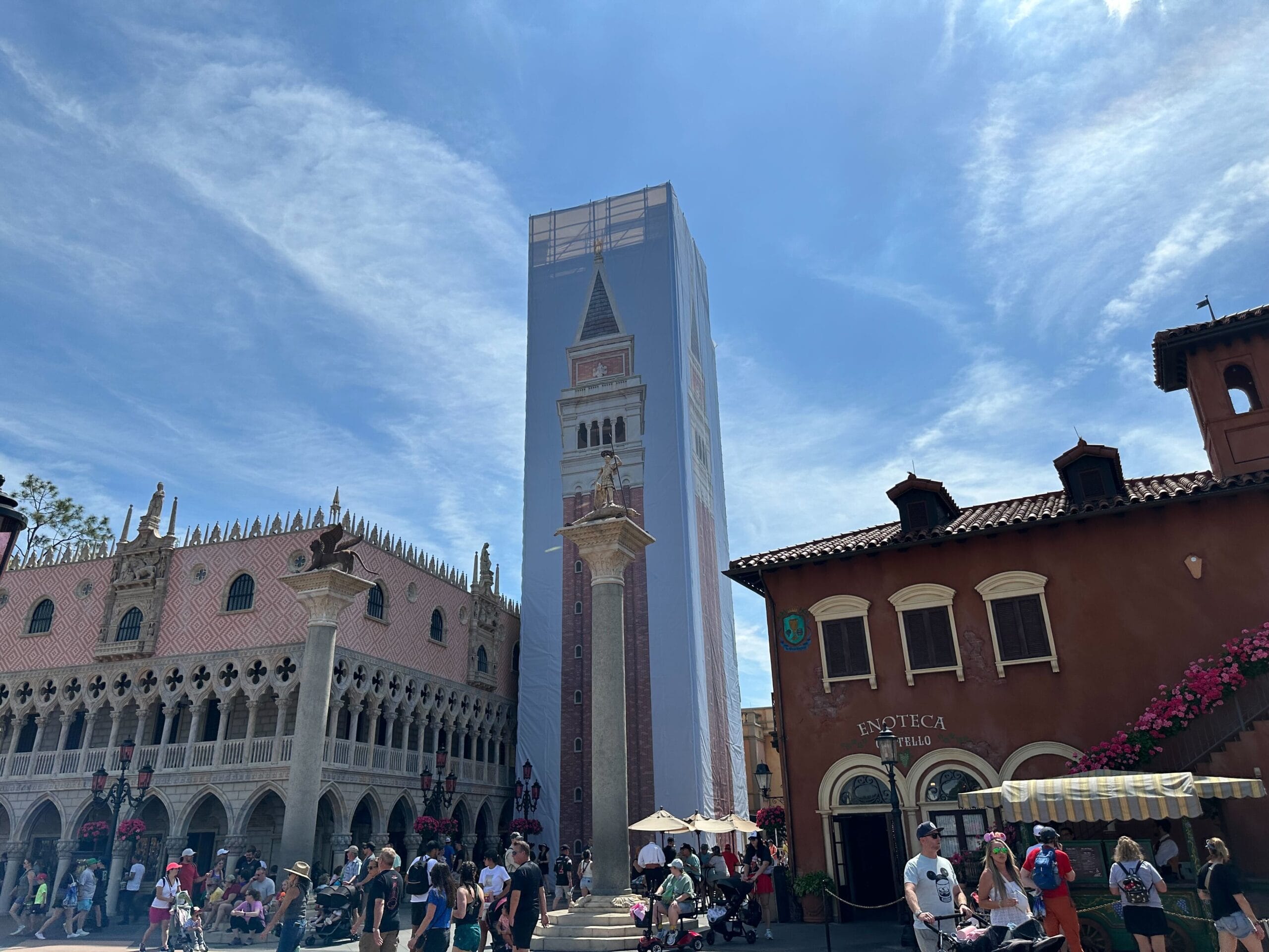 A tall building under construction, wrapped in a facade display featuring a bell tower image, stands surrounded by people and adjacent to European-style architecture, reminiscent of the Italy Pavilion at EPCOT, all under a clear blue sky.
