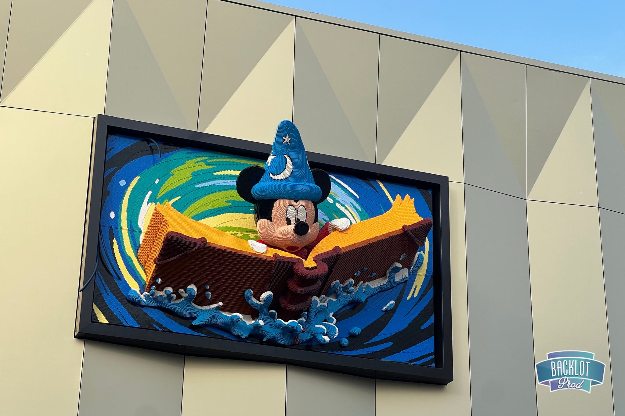 Wall art of a character resembling Sorcerer Mickey in a wizard hat riding a wave in a yellow boat. The artwork is mounted on a patterned wall with a "Backlot" sign in the corner, adding whimsical charm to the scene.