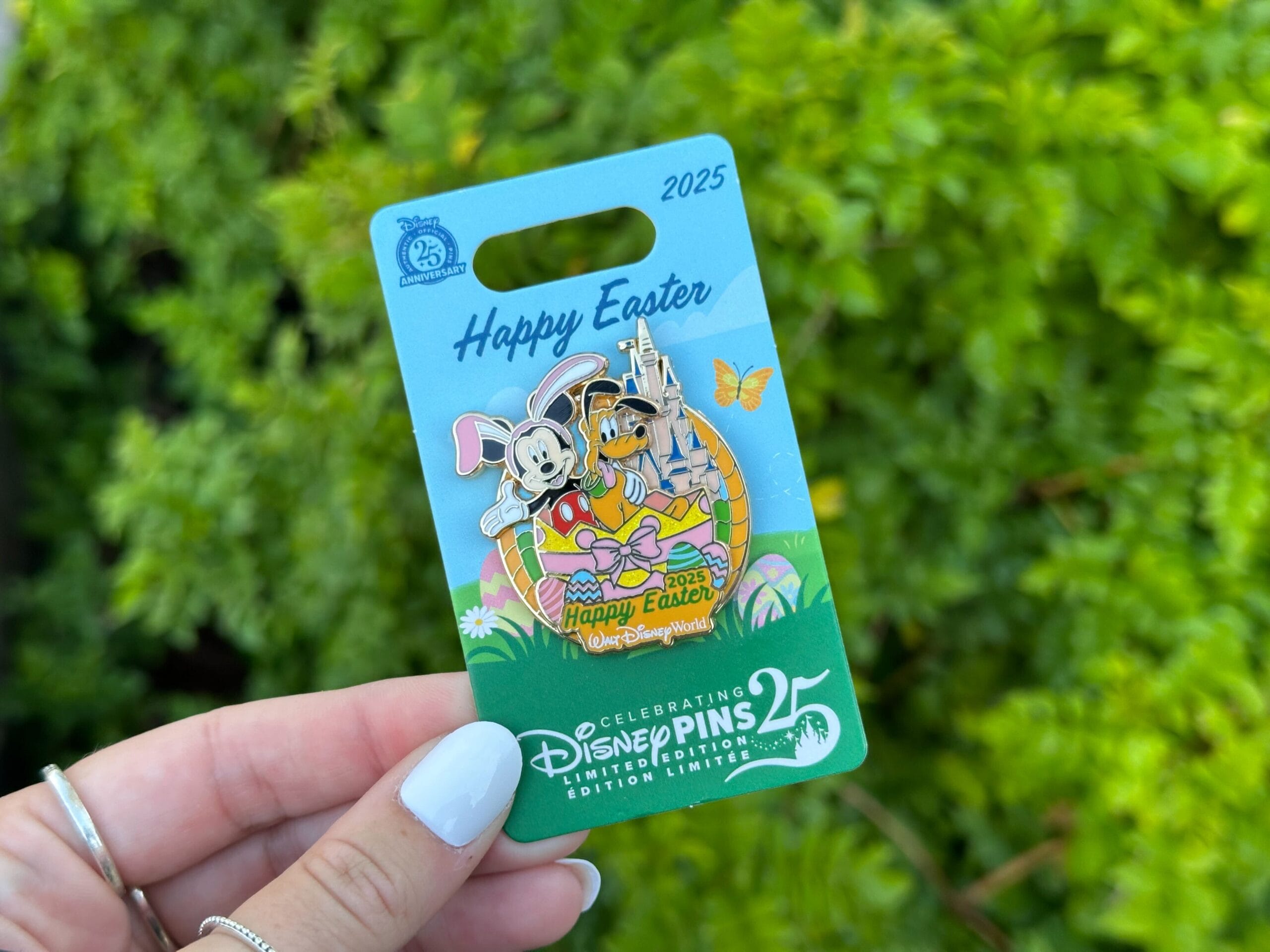 A hand holds a Disney Easter pin featuring Mickey and Minnie in bunny ears, with a Cinderella Castle illustration in the background. The pin is displayed on a themed card with greenery behind.
