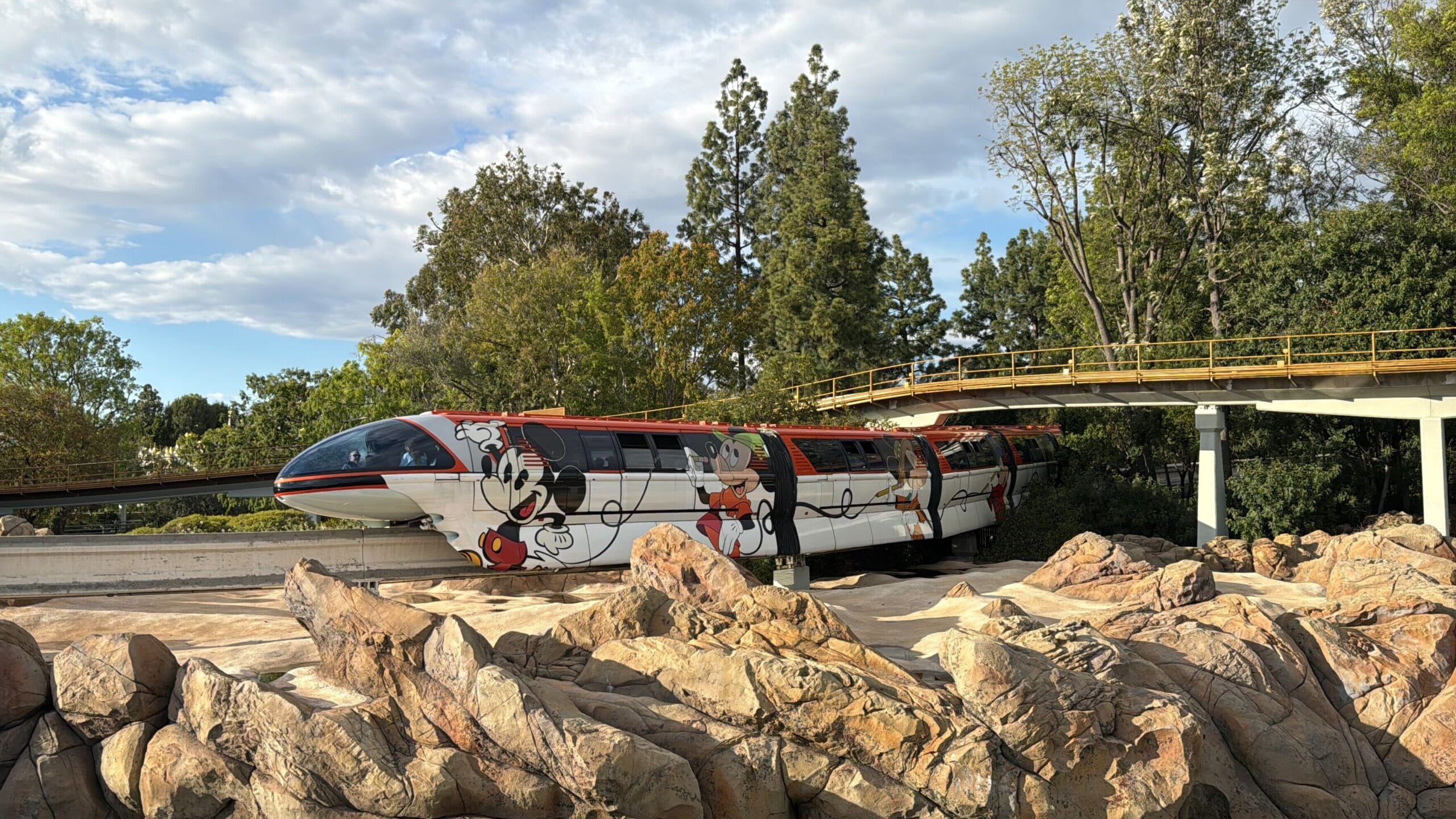 The Disneyland monorail, adorned with colorful cartoon character designs, glides along its reopened track, surrounded by lush trees and rocks beneath a partly cloudy sky.