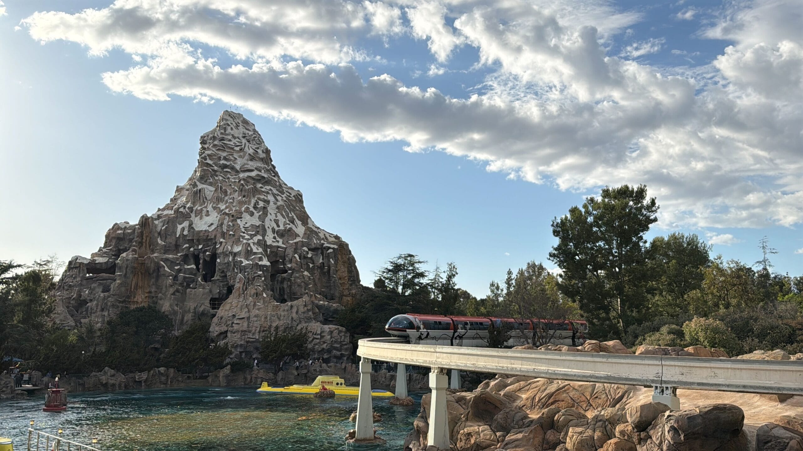 A red Disneyland Monorail travels on a raised track in front of a mountain with trees, while a submarine floats in the water below. The sky is partly cloudy, capturing the magic as it welcomes visitors once more.