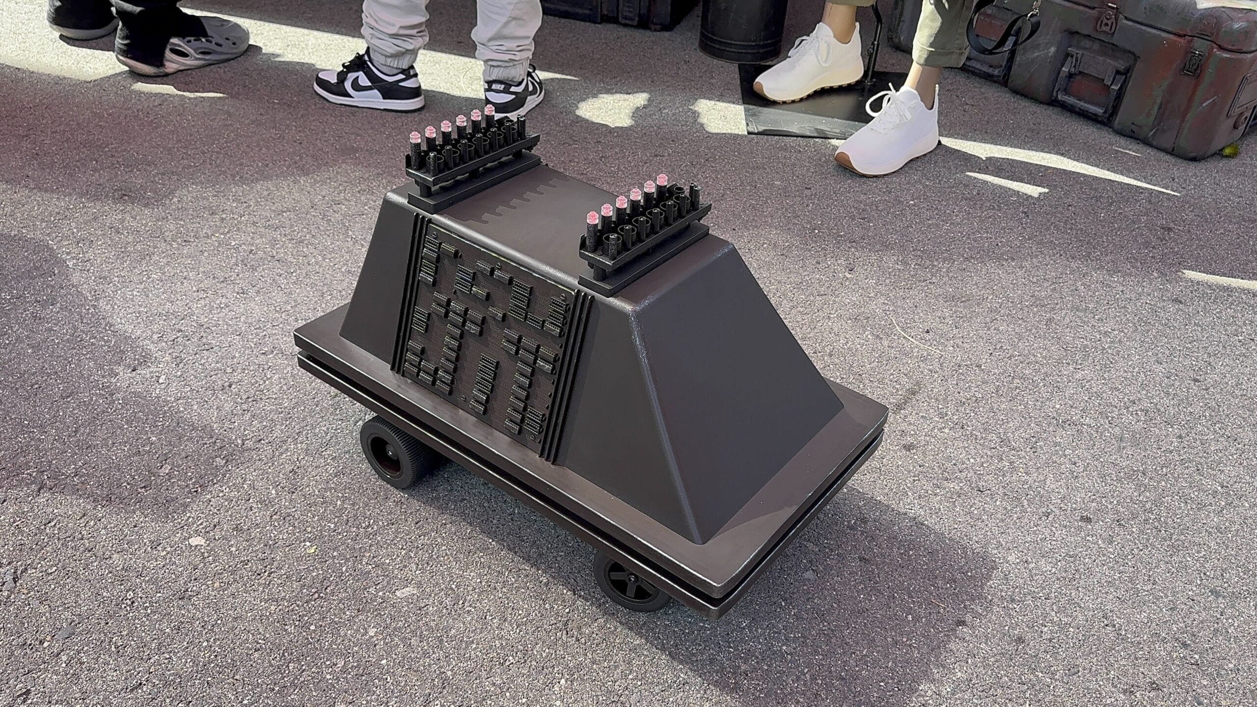 A sleek, RC Mouse Droid with a boxy, black design and two rows of pink-tipped spikes on top rests on the paved surface, resembling something out of a sci-fi VIDEO. People stand nearby, capturing PHOTOS of this intriguing, wheeled device.