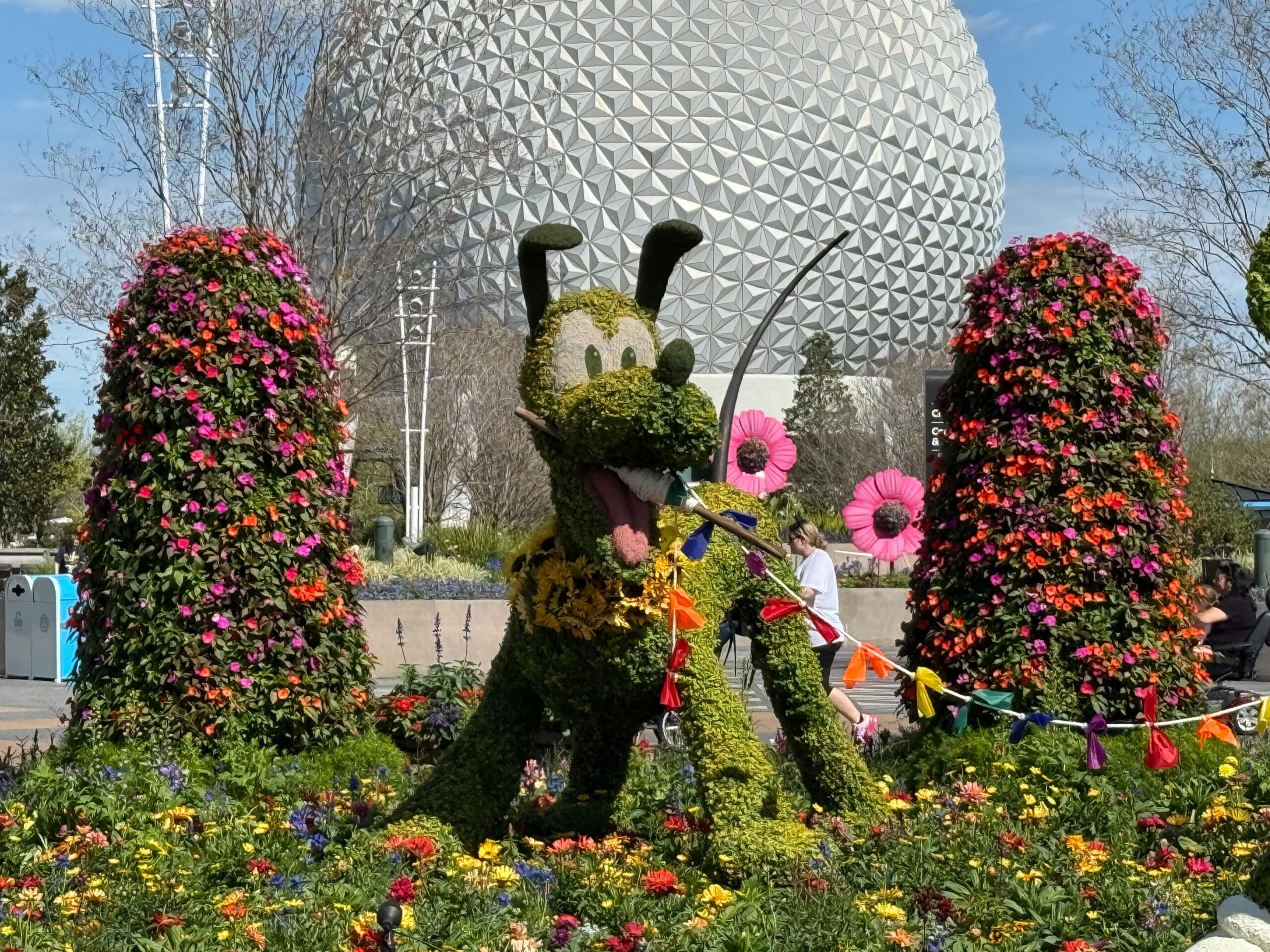 A whimsical topiary of a cartoon dog character sits amid vibrant flowers and plants, with a large geodesic dome structure rising in the background. The artistry of these playful topiaries brings the scene to life.