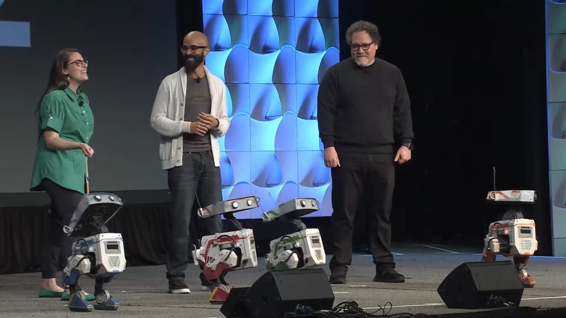 Three people stand on stage with three small BDX Droids, reminiscent of The Mandalorian's futuristic universe. The background features a geometric pattern, adding to the scene's sci-fi charm similar to a Disney Parks attraction.