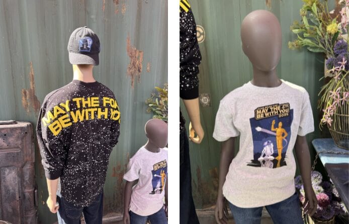 Two mannequins showcase "May the Force Be With You" shirts; one in a black sweatshirt and cap, the other in a white T-shirt. Positioned against a green wall, they offer a FIRST LOOK at Merchandise 2025 just in time for May the 4th Be With You celebrations.