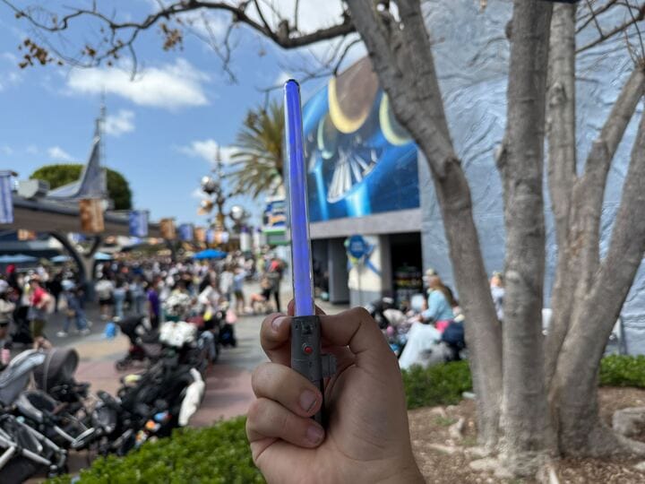 A person holds a blue Skywalker lightsaber in an outdoor area near a bustling theme park attraction at Disneyland, with strollers and eager visitors in the background.
