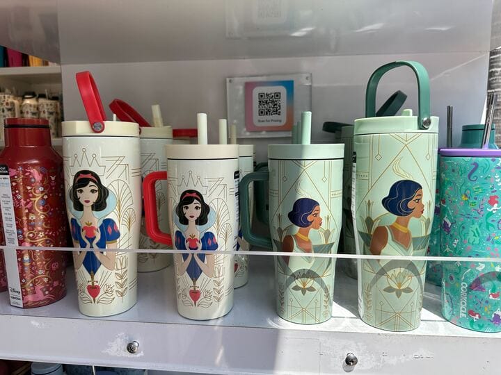 A display of Corkcicle Princess Tumblers featuring insulated water bottles with cartoon princess designs, some with red lids and others with green, reminiscent of a charming selection you'd find at Disney Springs.