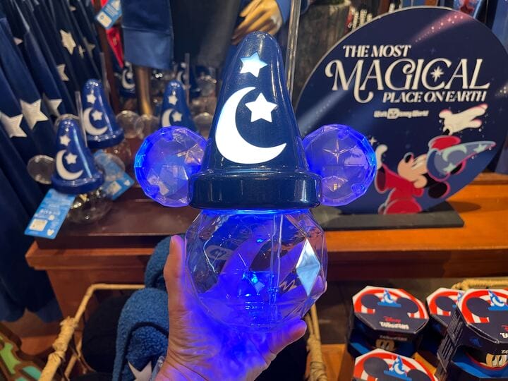 A hand delicately holds a glowing blue Mickey Mouse wizard hat light, embellished with stars and a moon, part of the enchanting Sorcerer Mickey Collection. The store brims with similar magical merchandise, inviting fans to savor each new discovery.
