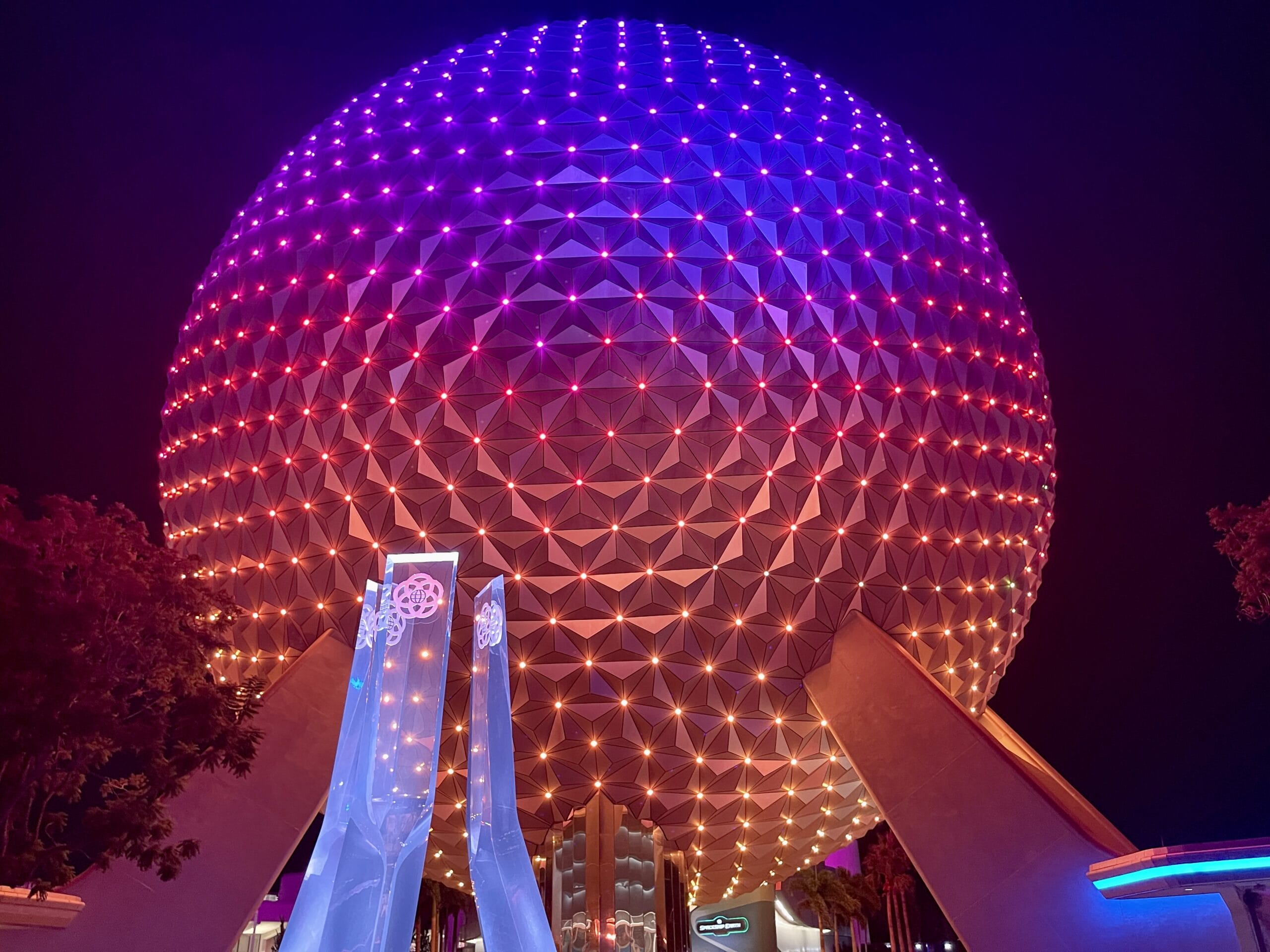 A geodesic sphere at EPCOT glows with purple and orange lights amidst futuristic structures, with a sign in the foreground capturing the night’s enchantment.