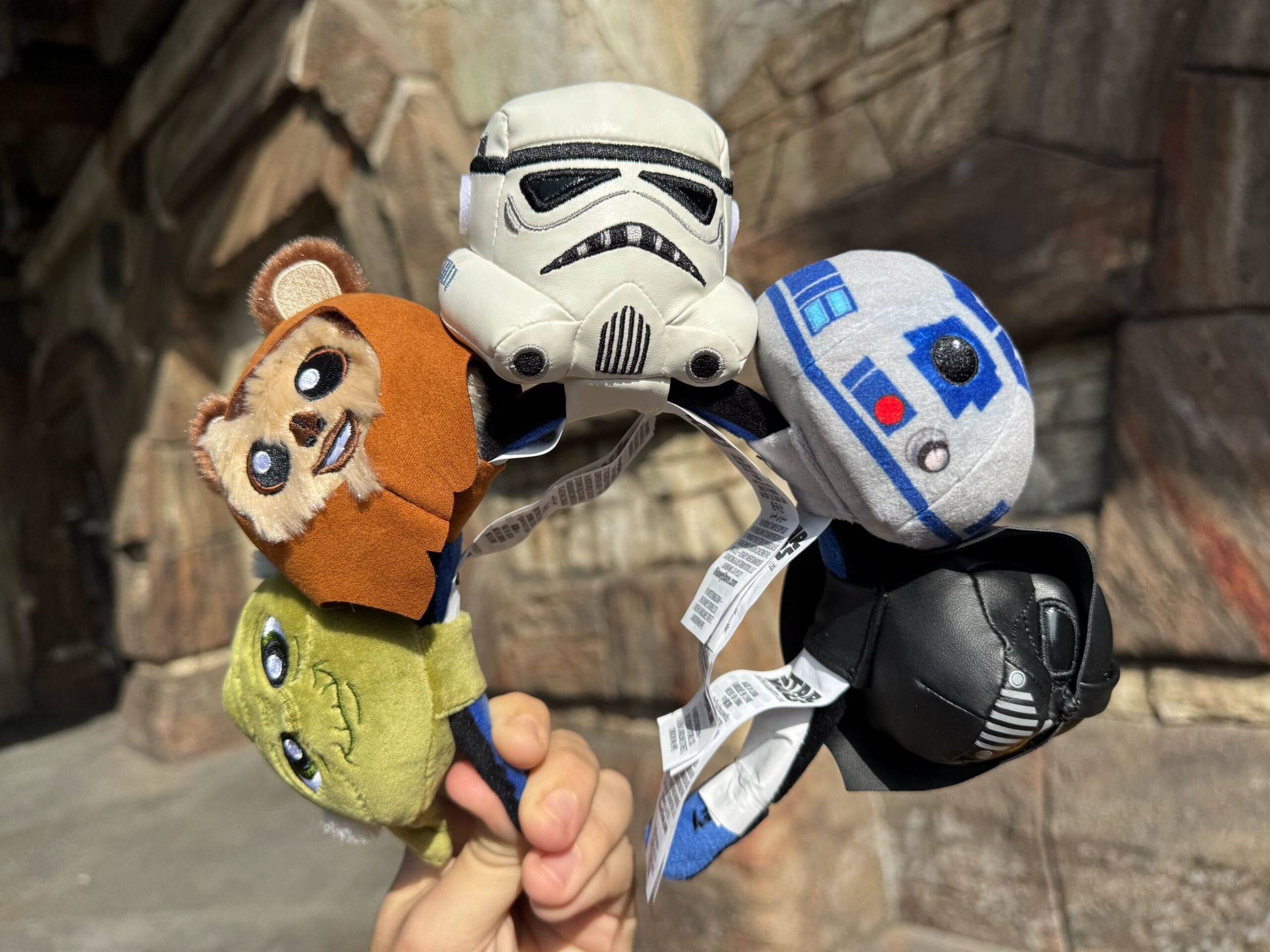 A hand showcases five Star Wars character plush toys on clips—an Ewok, Stormtrooper, R2-D2, Yoda, and Darth Vader—against a stone background. Perfect as a Disneyland headband plush collection for fans of all ages!.