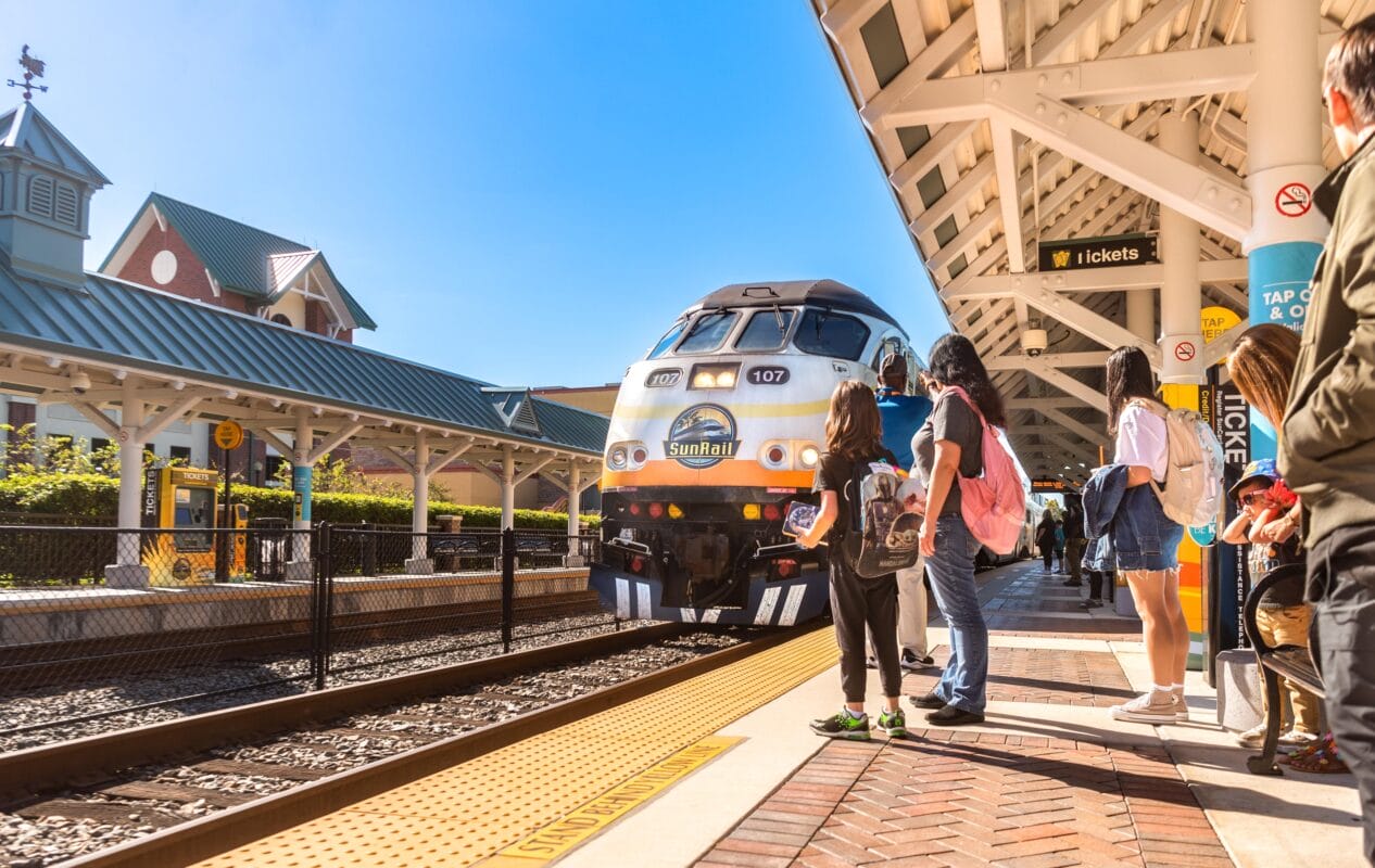 People eagerly wait on a platform as a SunRail train approaches, showcasing the vibrant Orlando transit system. The scene, bathed in sunlight and framed by a clear blue sky, highlights the charm of Central Florida.
