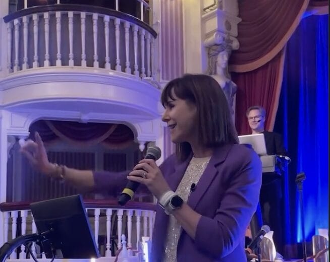 A woman in a purple blazer channels her inner Susan Egan, speaking into a microphone on stage, gesturing passionately. Behind her stands someone by a laptop, ready to cue the magic of 90s Nite. A balcony and draped curtains add to the nostalgic ambiance.
