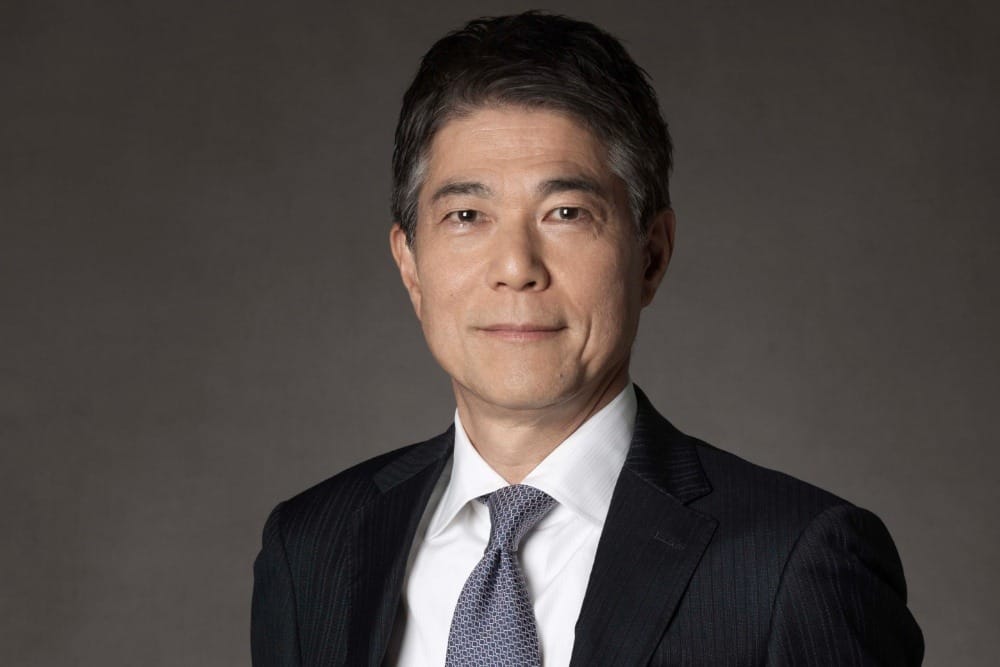 A person in a dark suit and tie stands against a plain gray background, looking at the camera with a neutral expression, evoking the understated elegance often seen in Disney's classic portrayals.