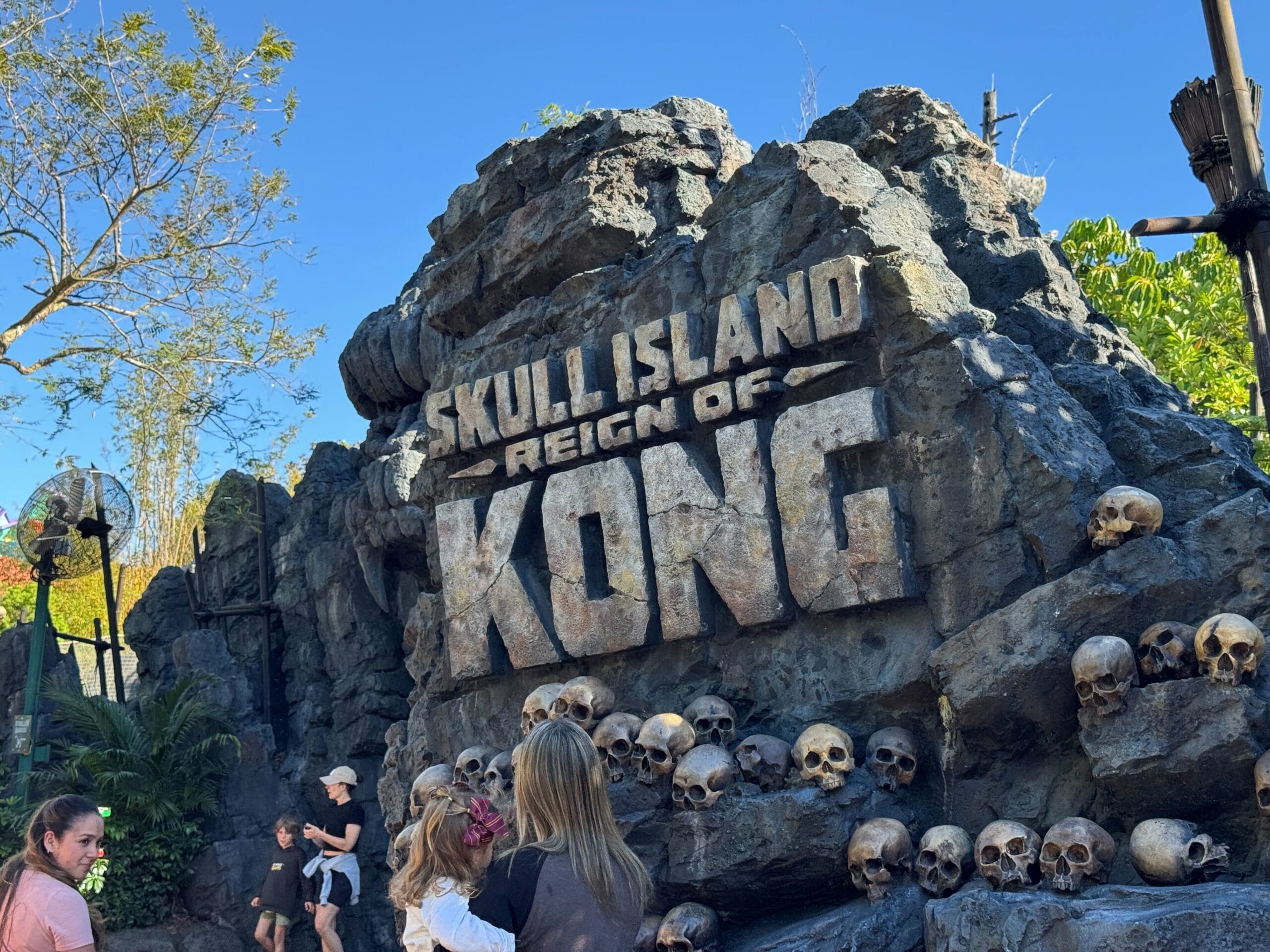 Visitors gather at the stone entrance of "Skull Island: Reign of Kong" at Universal Orlando Resort. Surrounded by ominous skulls and lush greenery, the scene unfolds underneath a clear, blue sky, promising an adventure like no other.