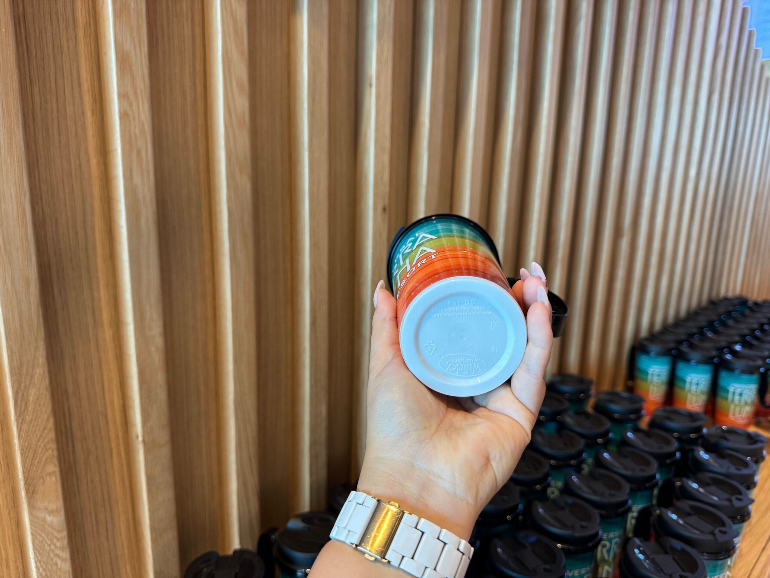 A hand holds a teal and orange mug with a Trevi logo, in front of a wooden slat wall. More identical mugs are stacked on the right.