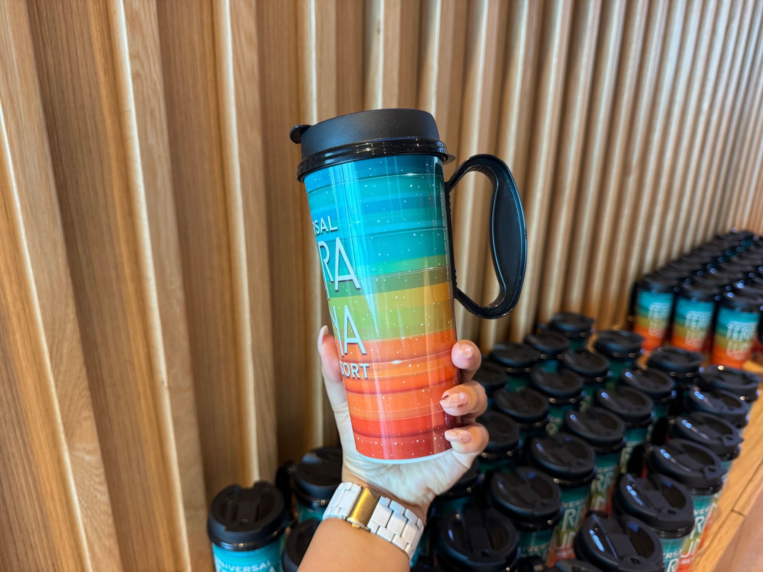 A person holds a colorful travel mug with a lid and handle in front of a wall lined with identical mugs.