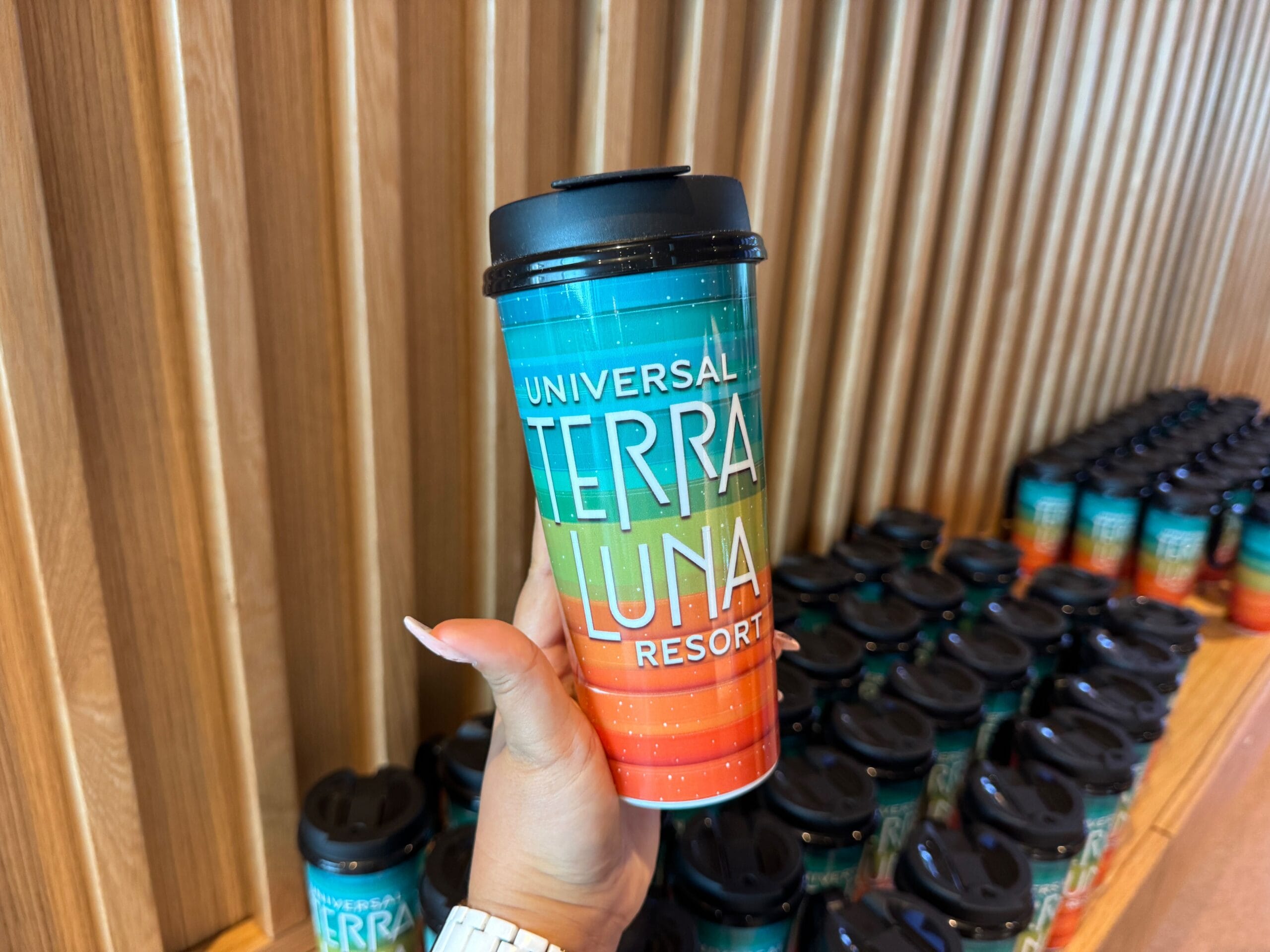 Hand holding a colorful travel mug with "Universal Terra Luna Resort" text, surrounded by more identical mugs on a wooden shelf.