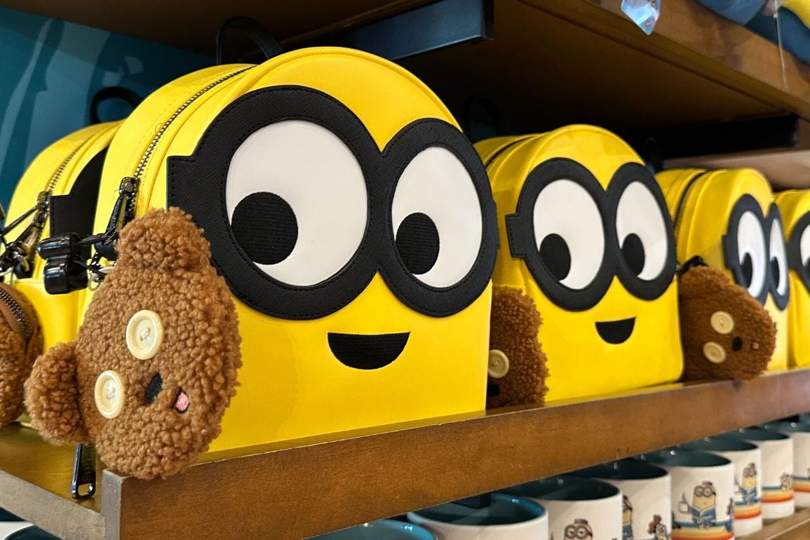 Yellow backpacks resembling cartoon characters, much like the playful Minion Loungefly Backpack, with large eyes and smiling faces are displayed on a shelf. Each backpack boasts a brown plush accessory attached, adding a touch of whimsy.