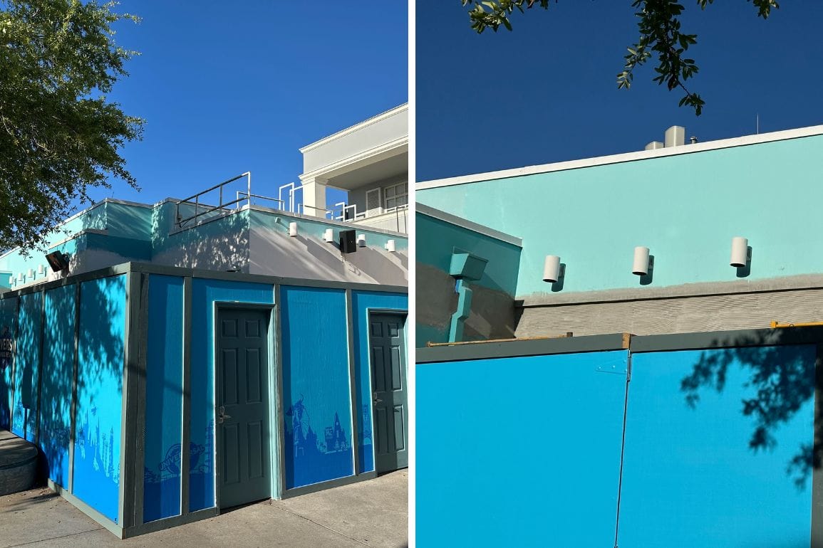 A building with bright blue walls and a flat roof, partially obscured by construction barriers, stands shaded by nearby trees under a clear blue sky. This On Location shop is a vibrant spot in the midst of its transformation.