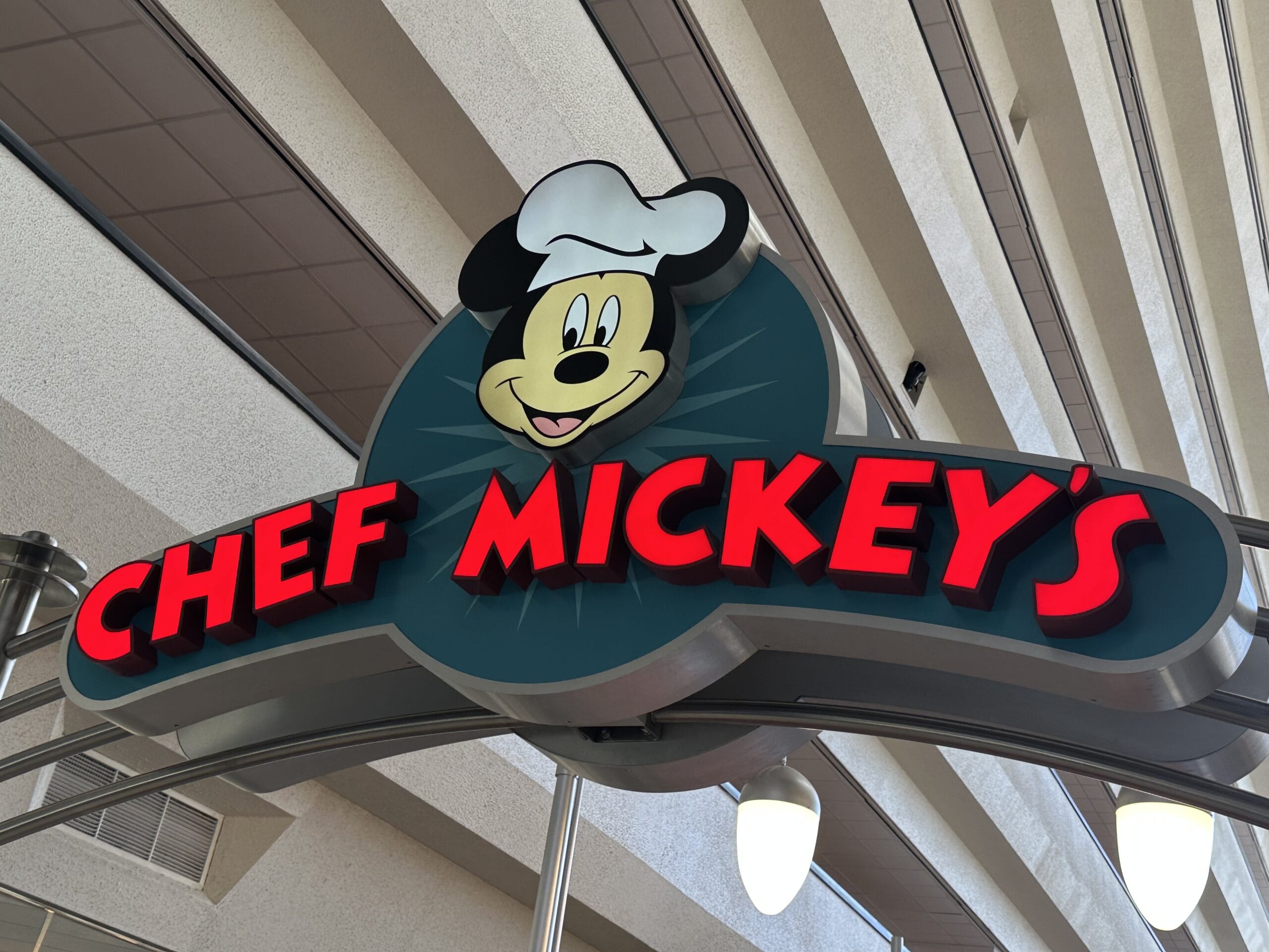 A whimsical sign for "Chef Mickey's" featuring a cheerful Mickey Mouse in a chef's hat, set against a ceiling with sleek linear architectural elements, captures the playful essence of Pop Century.