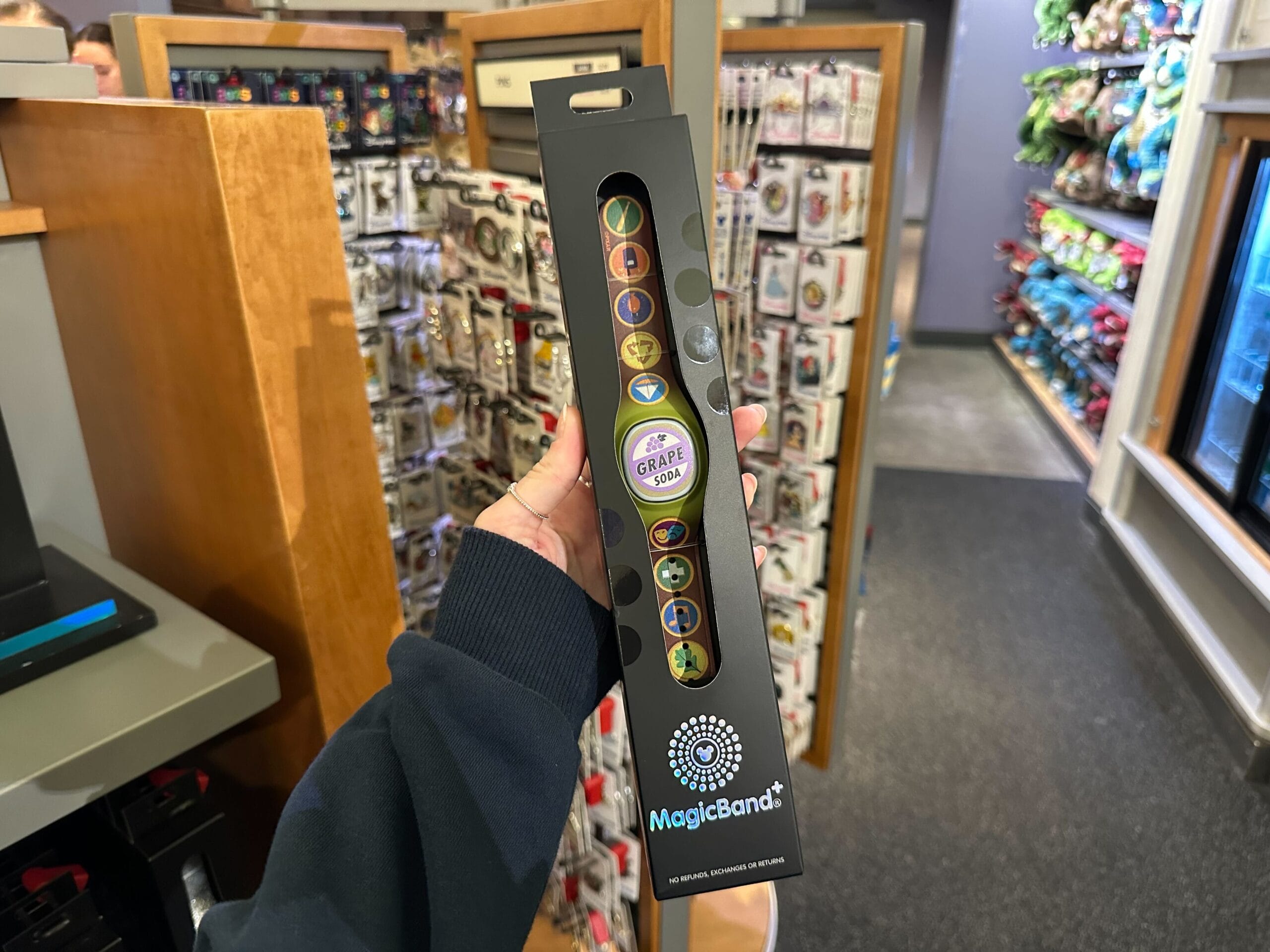 Hand holding a boxed MagicBand+ themed with Figment in a store, surrounded by various merchandise.