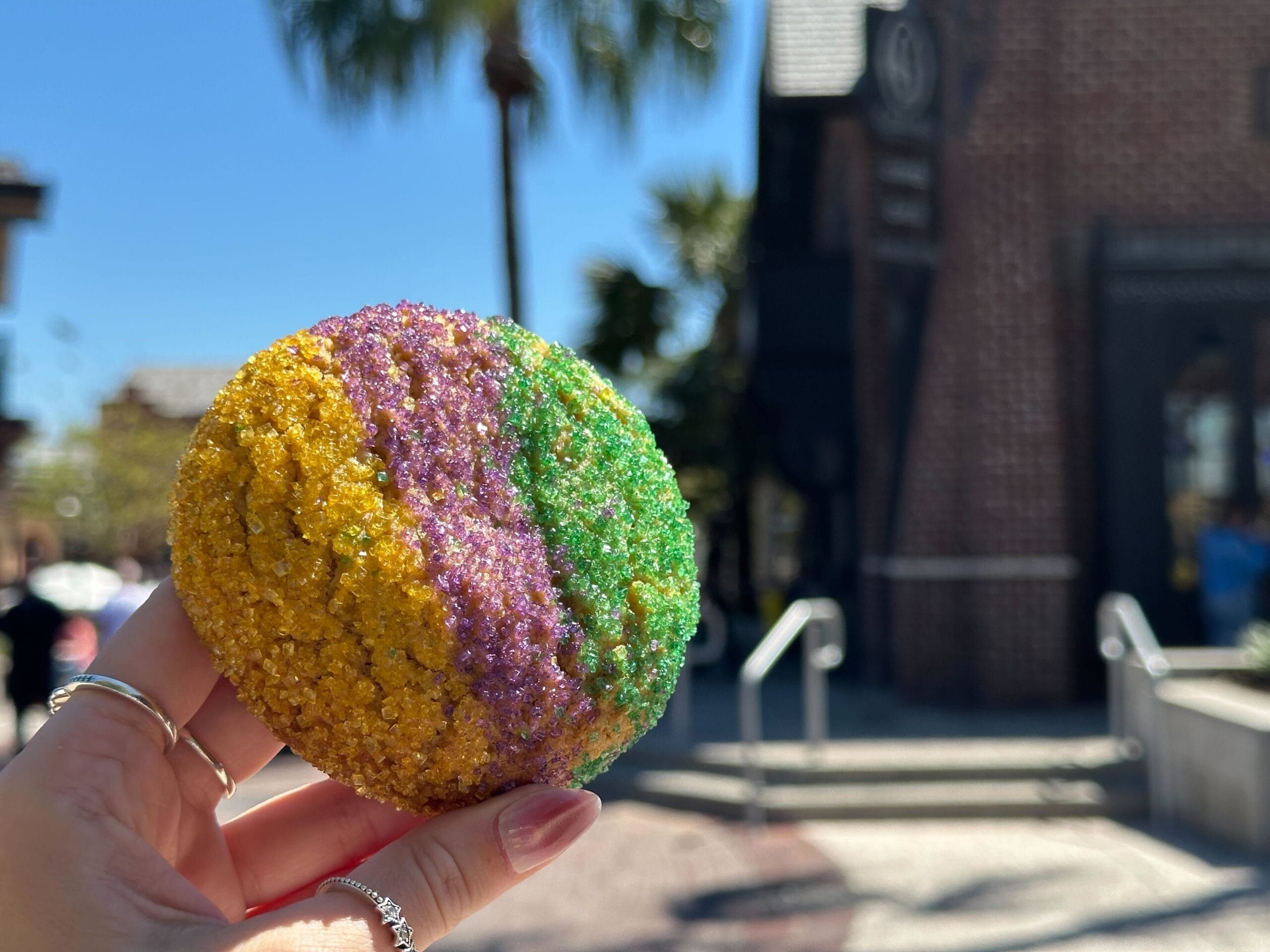 A hand holds a King Cake Sugar Cookie with yellow, purple, and green stripes against an urban backdrop of buildings and palm trees, reminiscent of the artistry seen at Gideon's Bakehouse in Disney Springs.