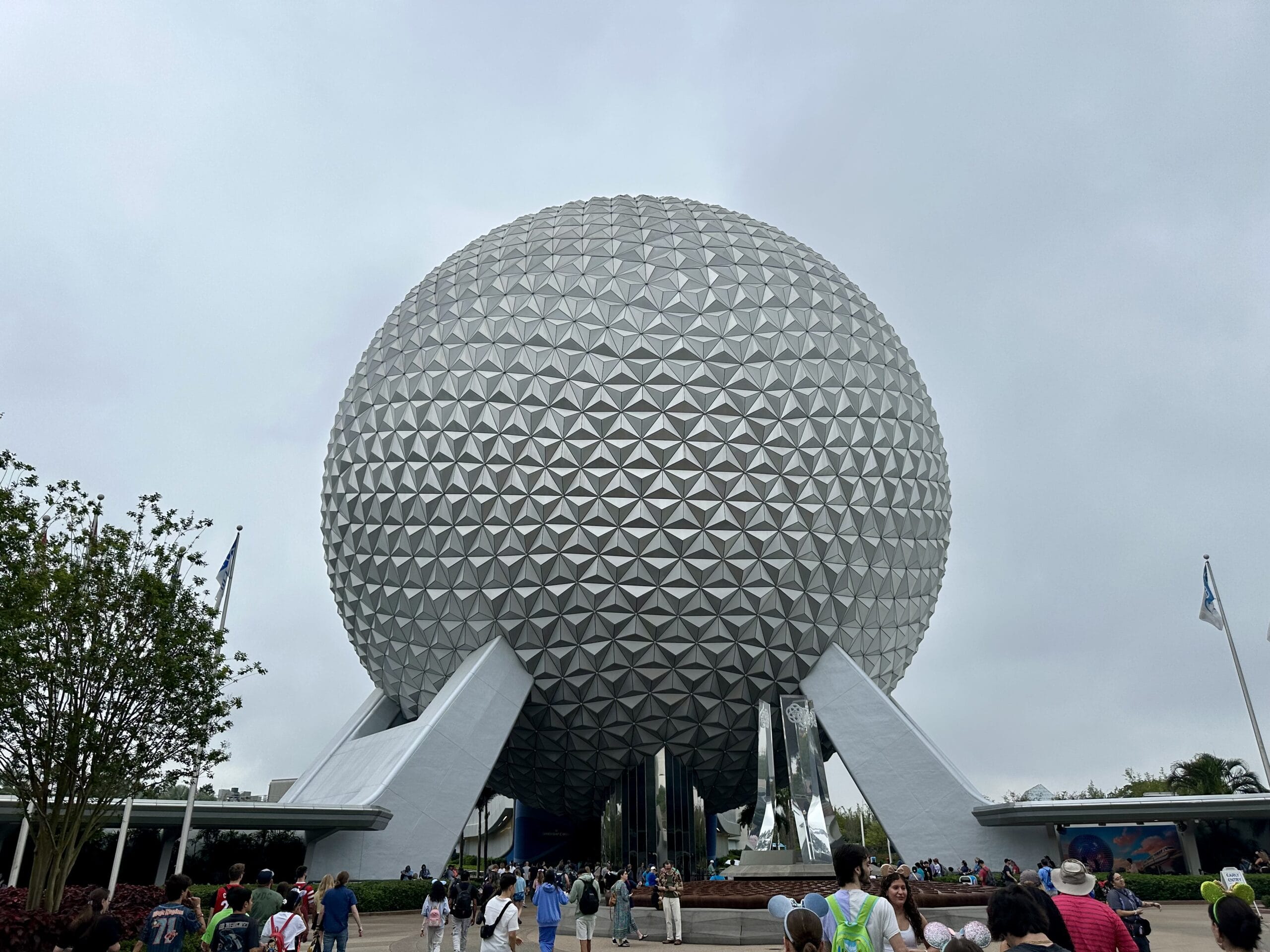 A large geodesic sphere with a geometric pattern stands grandly on stilts, surrounded by people at its base in EPCOT. An overcast sky looms in the background, adding to the mystical allure of this architectural marvel.