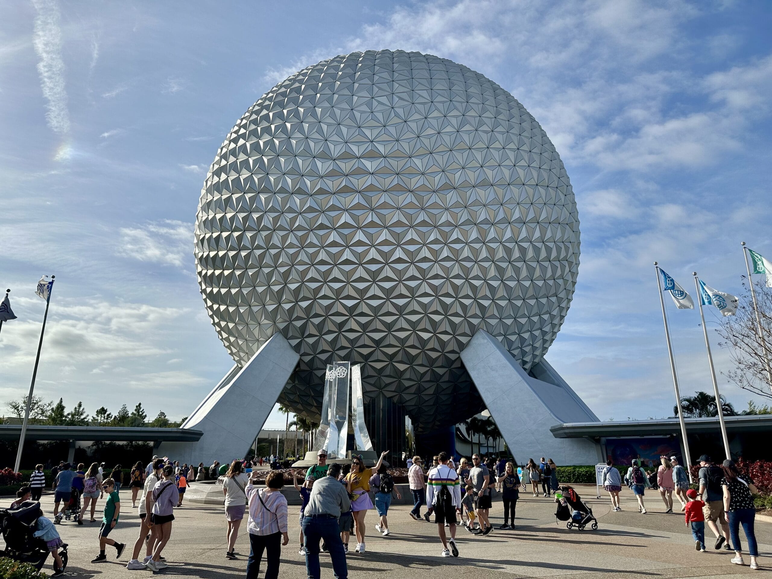 A large geodesic sphere with triangular panels stands prominently at EPCOT, echoing the innovation of Disney World. People stroll under a clear blue sky, surrounded by vibrant Flower and Garden displays. Flags flutter to the right, completing the picturesque scene.