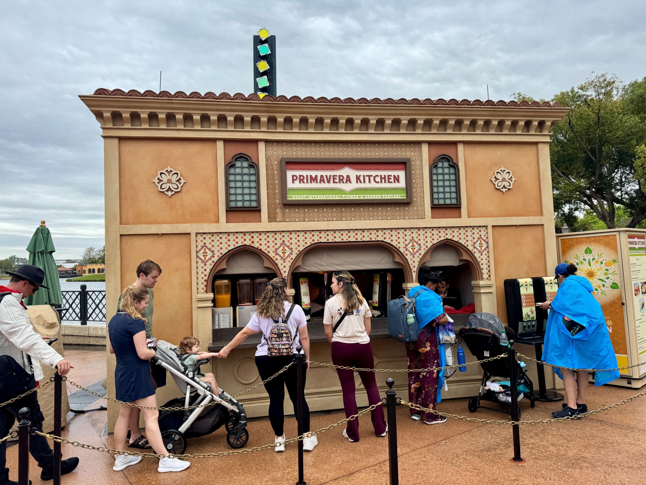 People line up at Primavera Kitchen, a food stand with a brown and orange facade adorned with Italian elements. As part of the Flower & Garden Festival, the lively scene includes families with strollers and colorful umbrellas scattered around.