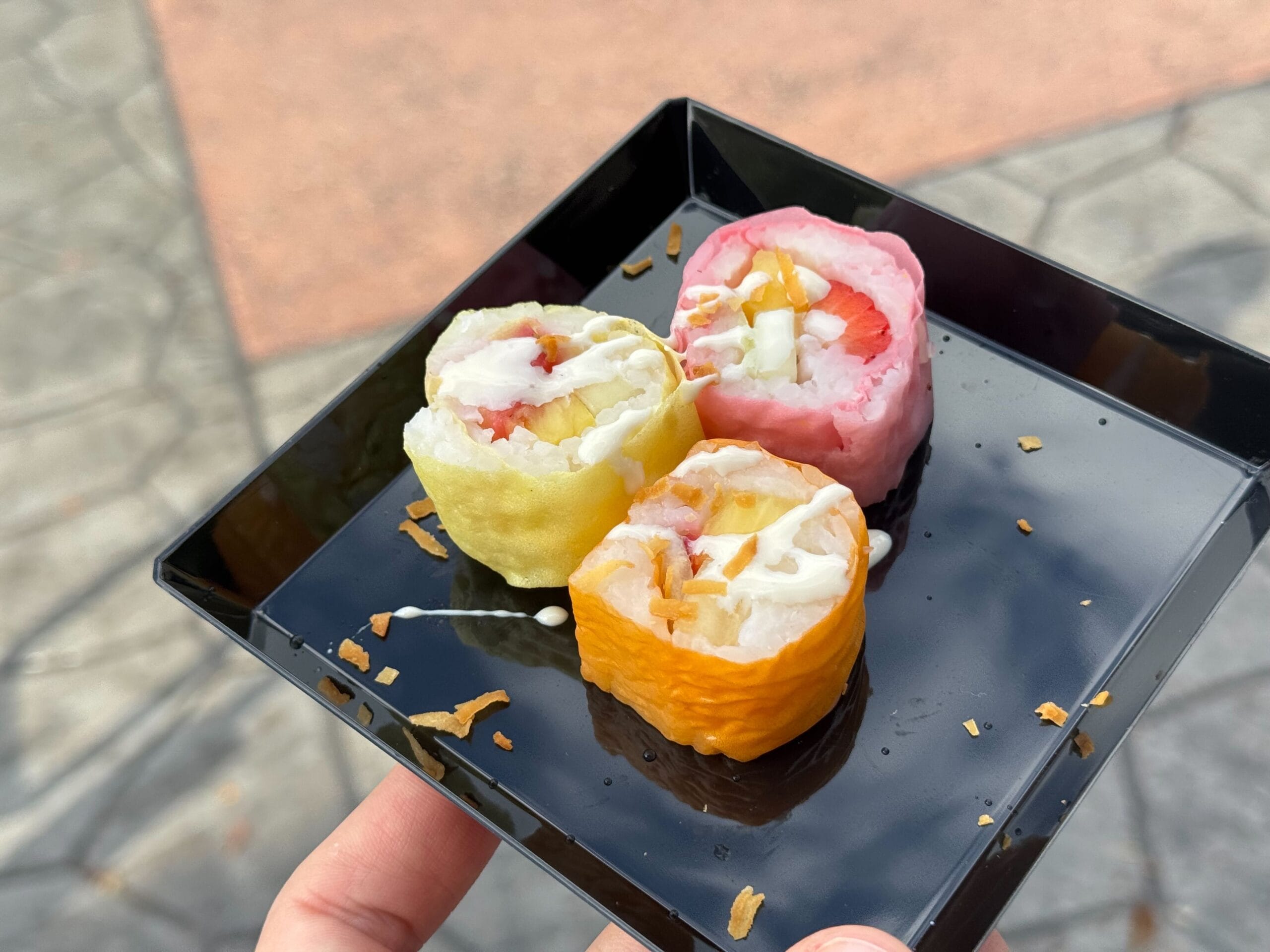 Three colorful sushi rolls with creamy filling sit elegantly on a black square plate, ready to be enjoyed at the Flower & Garden Festival, beautifully blending culinary art with nature.