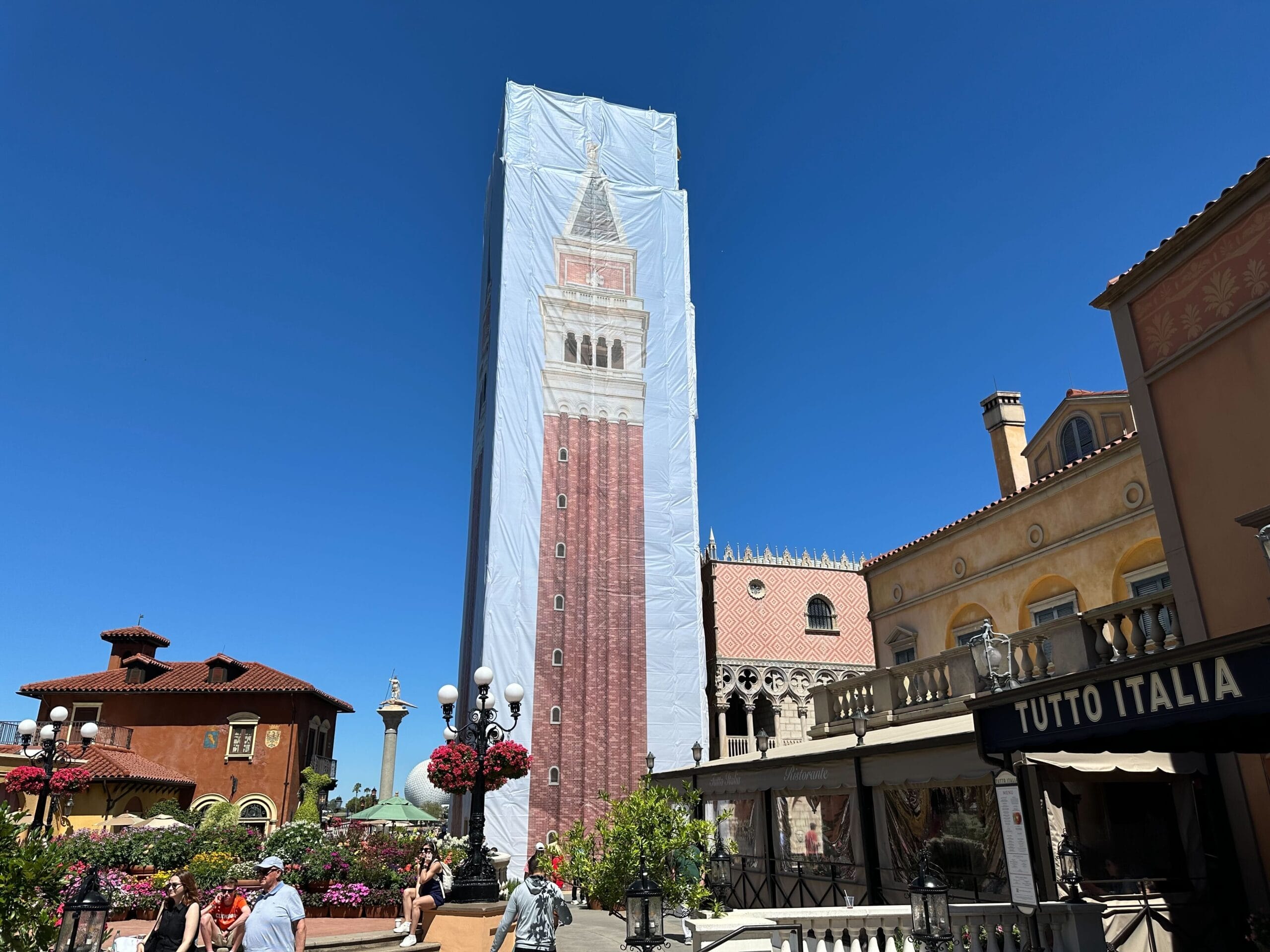 A building wrapped in a tarp with a tower design, under a clear blue sky. Surrounding it are outdoor lamps, potted flowers, and the facade of "Tutto Italia" restaurant.