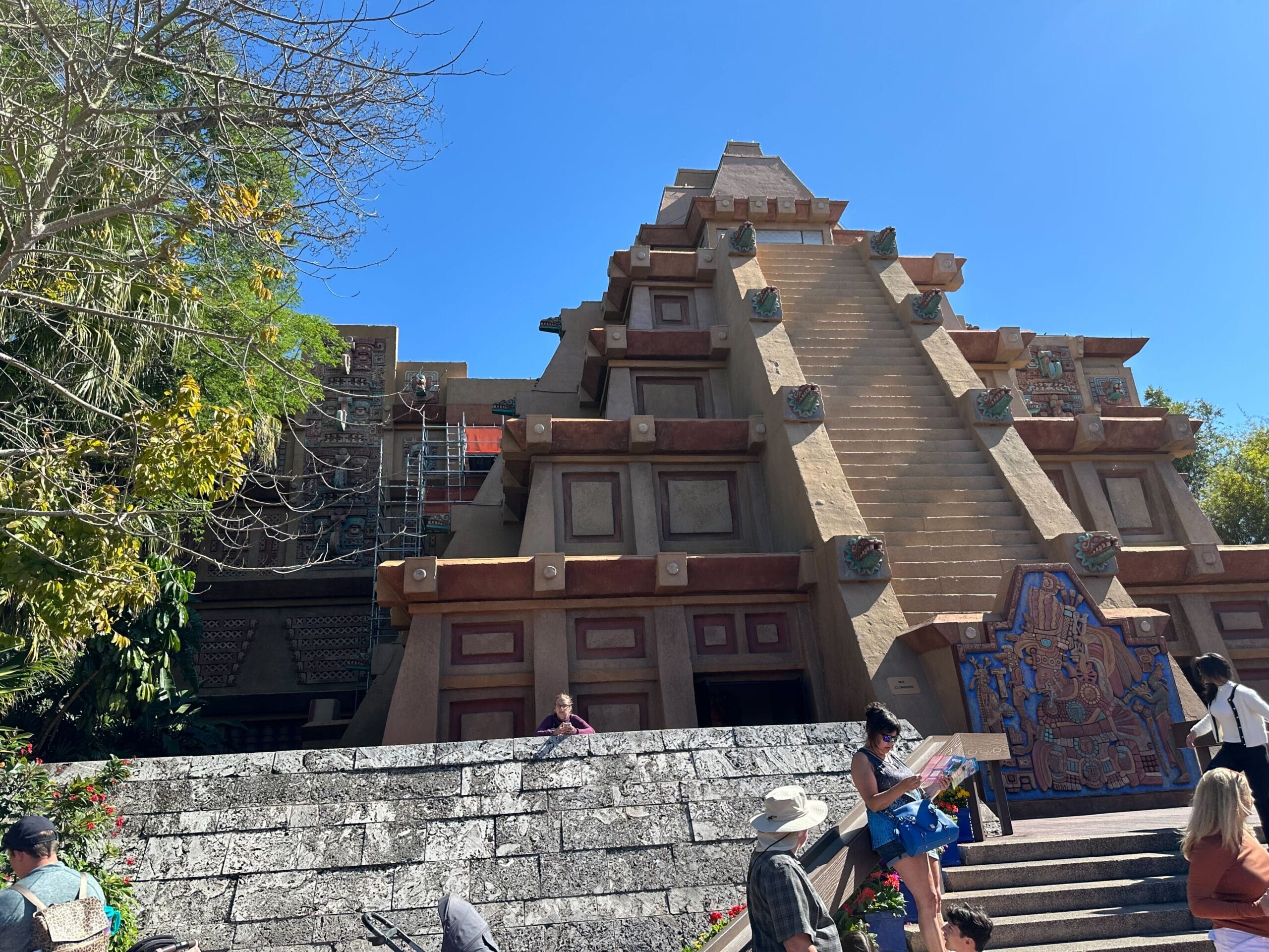 A pyramid structure with steps, adorned with colorful carvings, under a clear blue sky. People stand and walk around the base, surrounded by trees and a stone wall.