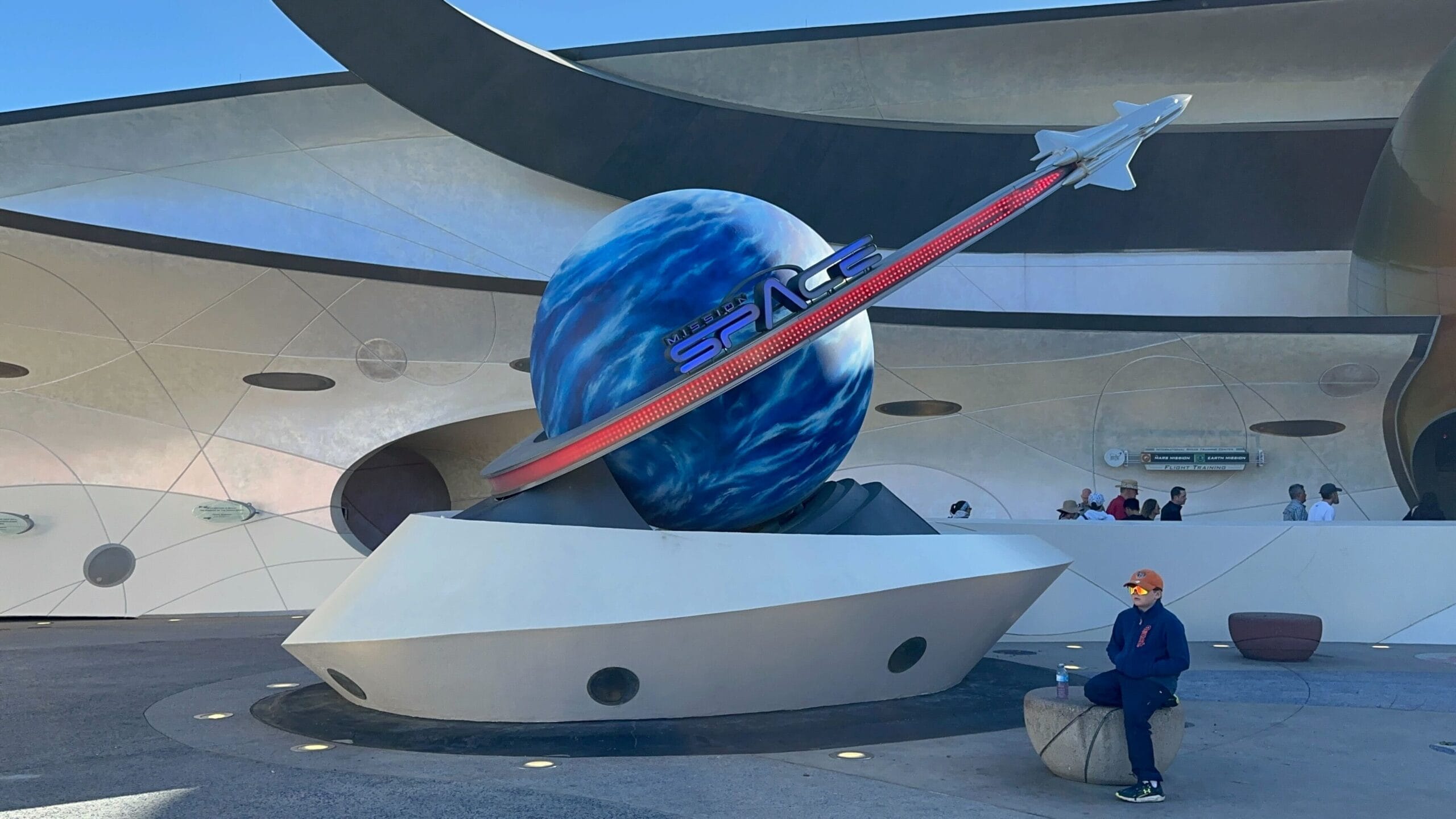 A person sits on a stone bench beside a futuristic sculpture featuring a blue planet with a soaring spacecraft and the word "Space.