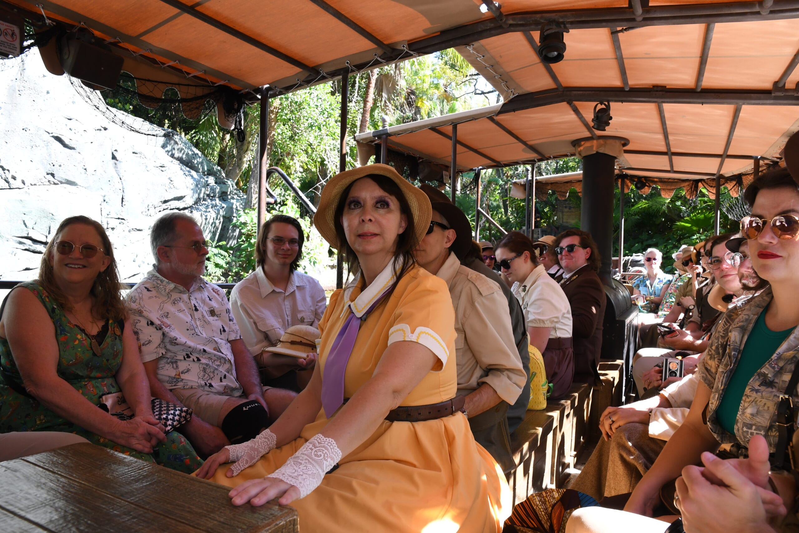 A group of people in vintage clothing sits on a boat ride with wooded seating and a canopy cover, surrounded by greenery; it's a whimsical scene straight out of Magic Kingdom's Adventureland Day.