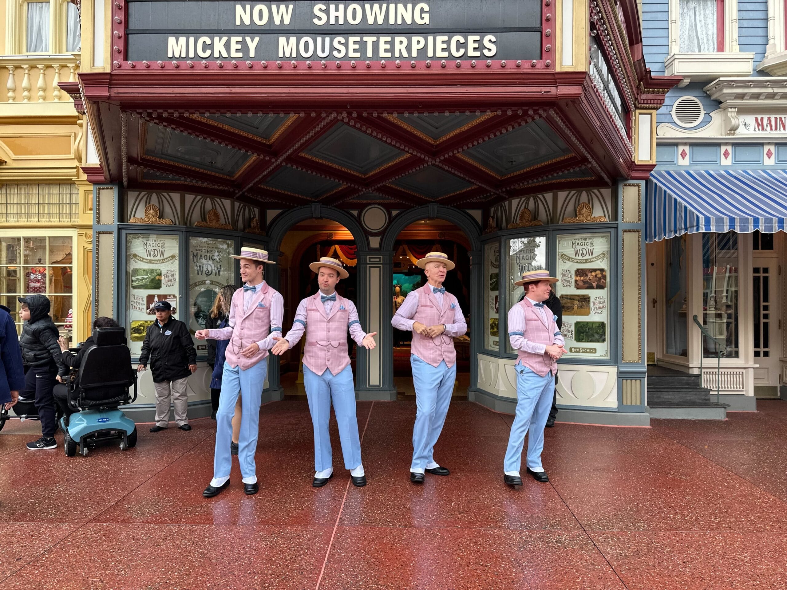 Four performers in pastel suits and hats sing outside a theater with "Mickey Mouseterpieces" on the marquee. Pedestrians pass by, and a person in a wheelchair observes.