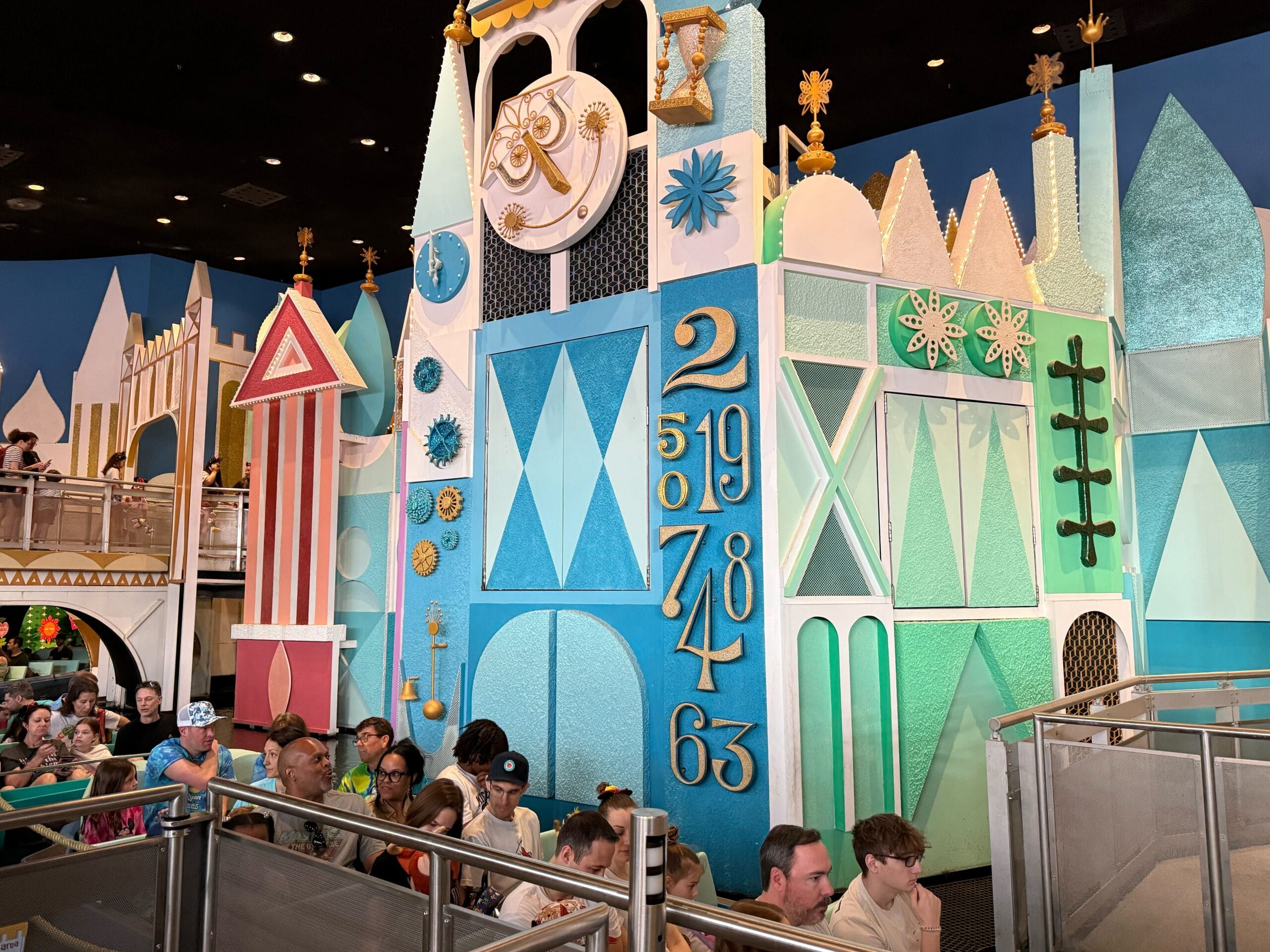 People wait in line inside a Magic Kingdom attraction adorned with colorful geometric designs and a whimsical clock tower, reminiscent of the charming style found in it's a small world.
