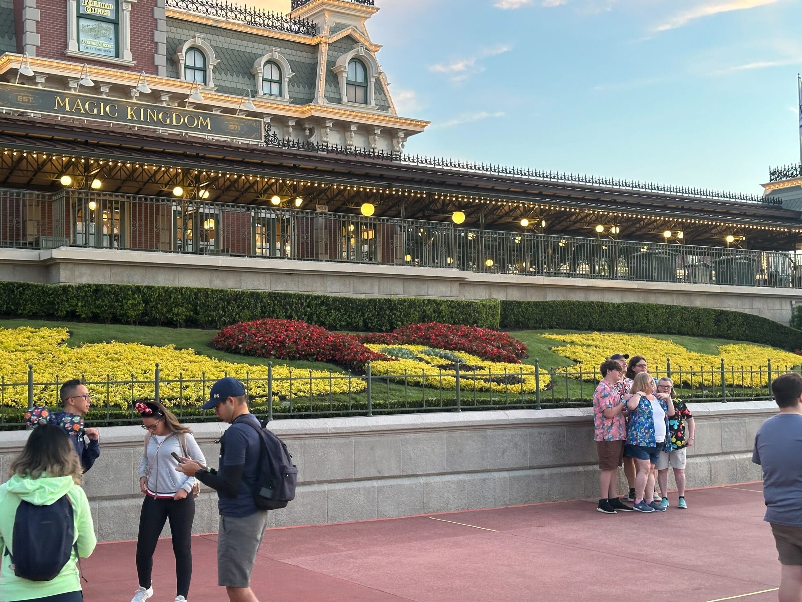 People stand and walk near the entrance of Magic Kingdom, featuring a floral display and vintage-style building with "Magic Kingdom" signage above.