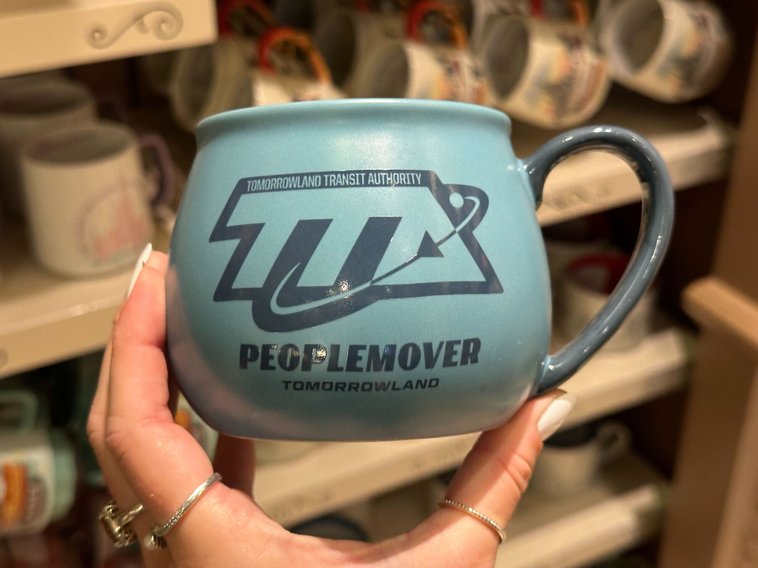 A blue mug with a Tomorrowland Transit Authority PeopleMover logo, featuring a stylized arrow and planet icon, is a delightful nod to Walt Disney World's iconic attraction.