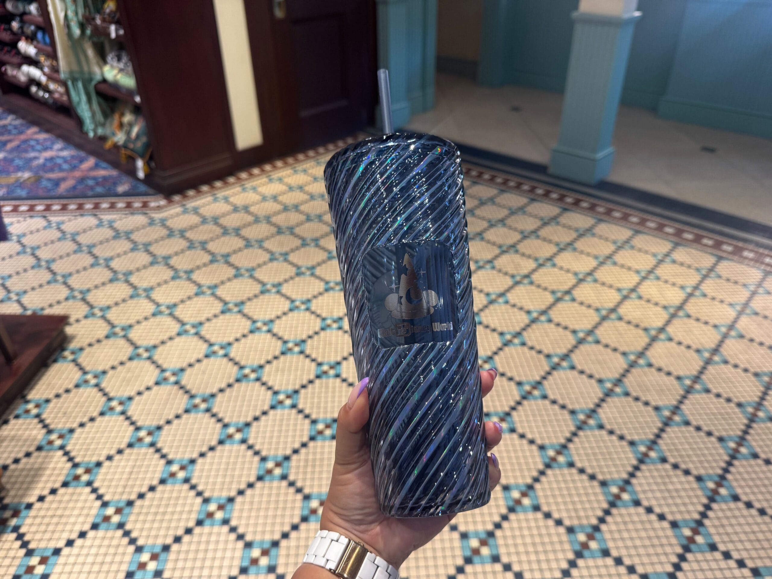 A hand holding a blue, intricately patterned tumbler with a straw inside a room with geometric tile flooring.