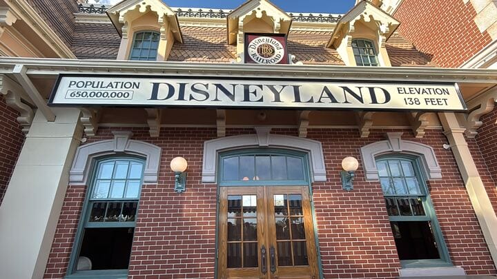 A brick building with a sign reading "DISNEYLAND Population 650,000,000 Elevation 138 feet" above the entrance stands proudly as the Disneyland Railroad reopens. The roof features dormer windows and decorative elements, echoing the charm of classic construction.
