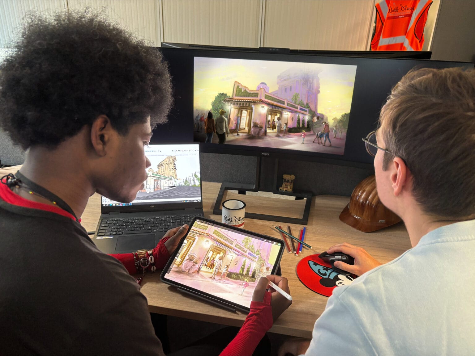 Two people work at a desk with a digital tablet and dual monitors displaying an architectural illustration. A hard hat and mug are nearby.