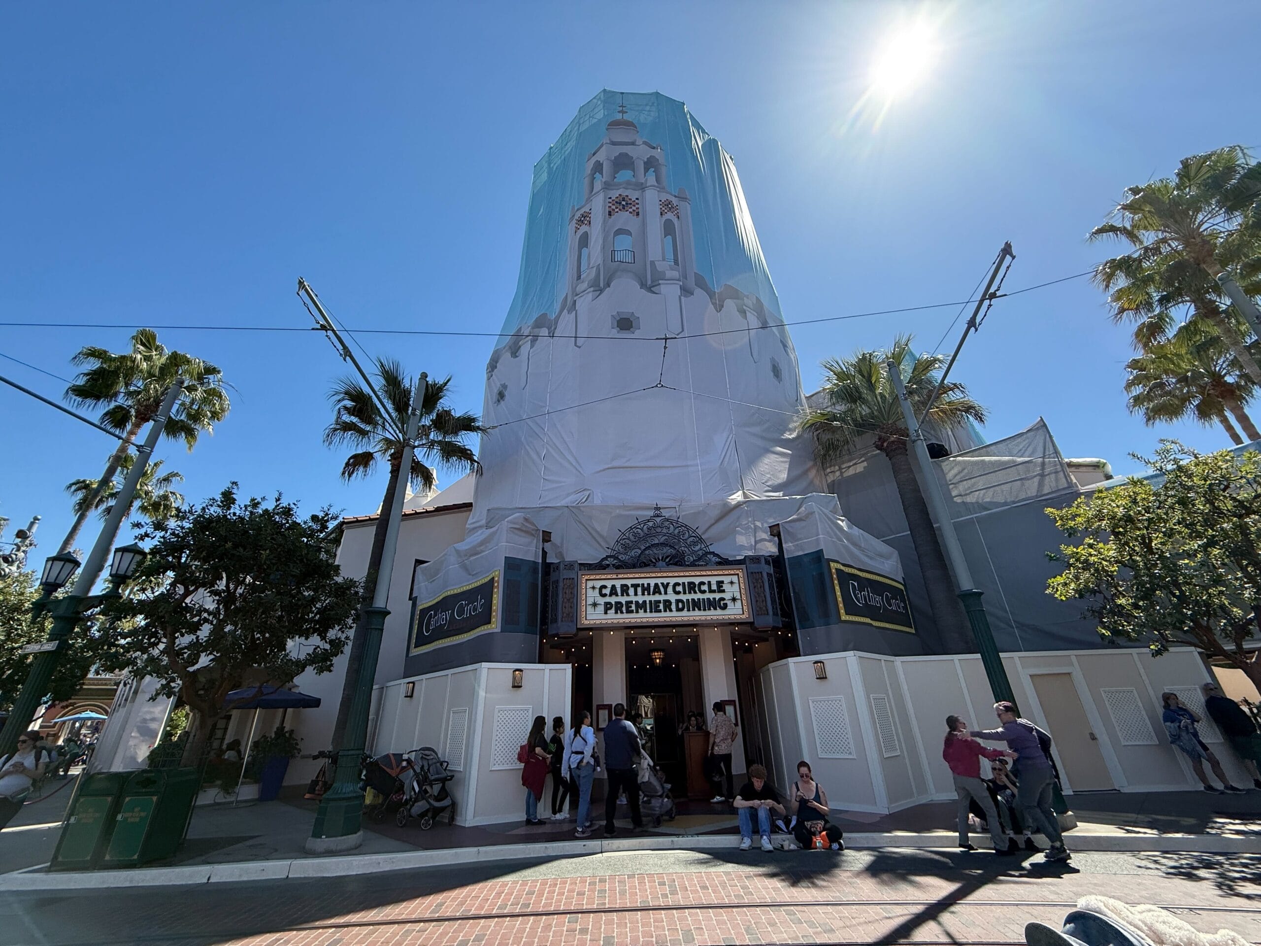 The Carthay Circle building under renovation, wrapped in scaffolding and cover. People are gathered at the entrance and sitting nearby. Sunny day with palm trees in the background.