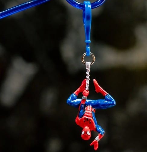 A small Spider-Man figure hangs upside down from a blue plastic hook, as if swinging through the streets of Disneyland Resort, holding onto a white string against a dark background.
