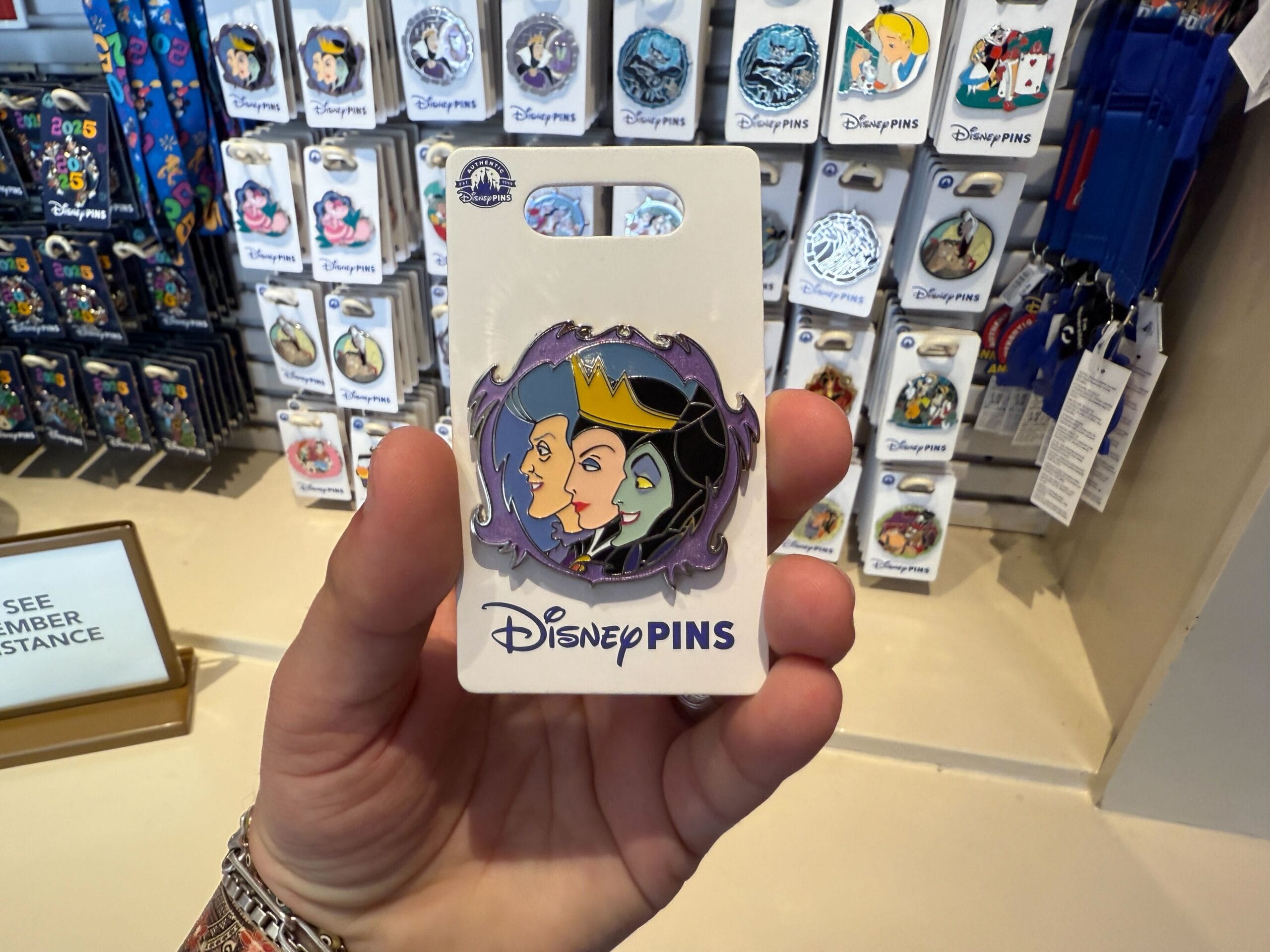 Person holding a Disney pin featuring three villainous characters in front of a display of various other Disney pins.