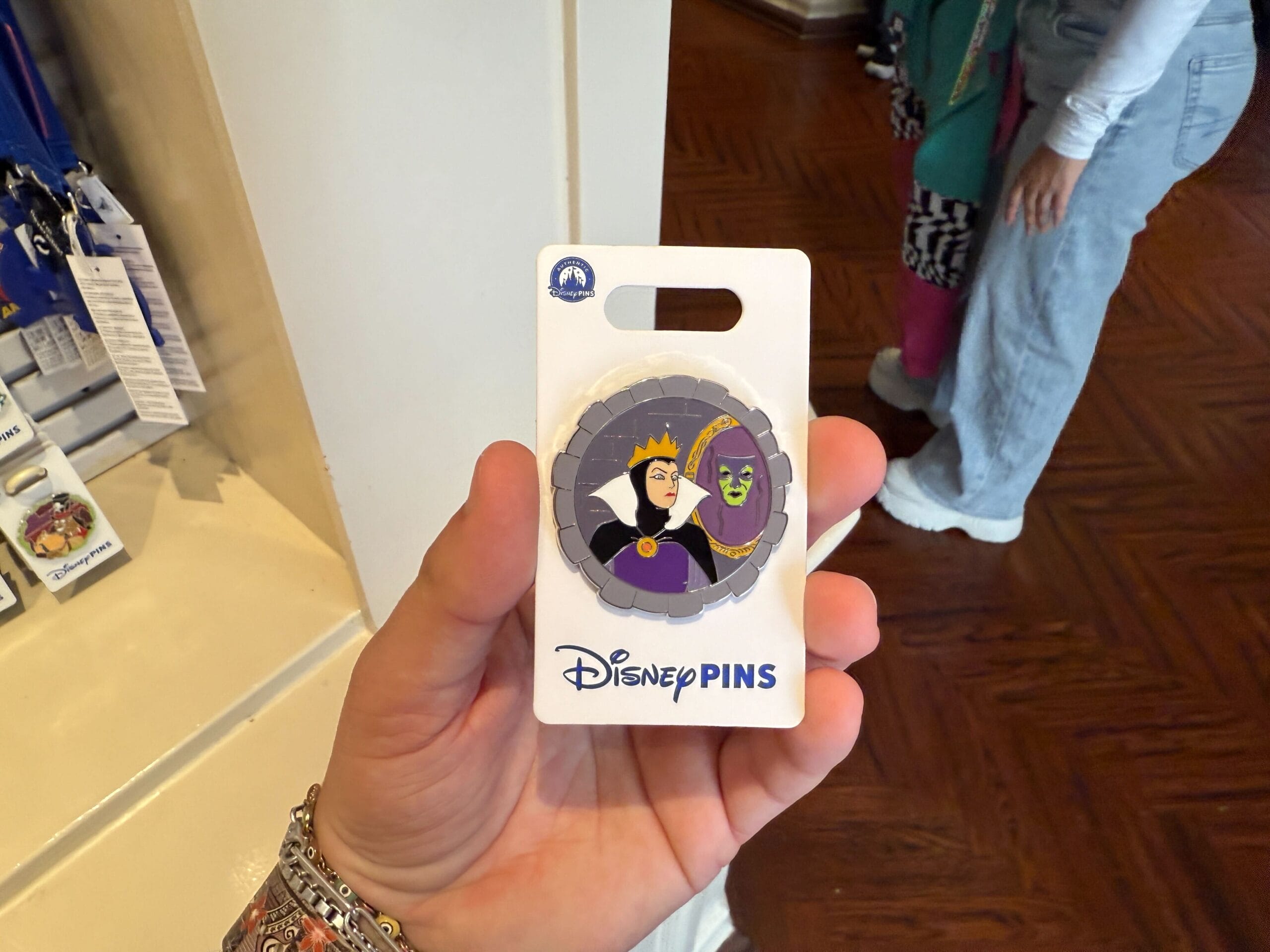 Hand holding a Disney pin featuring the Evil Queen from Snow White with a mirror. Other pins and items visible in the background.