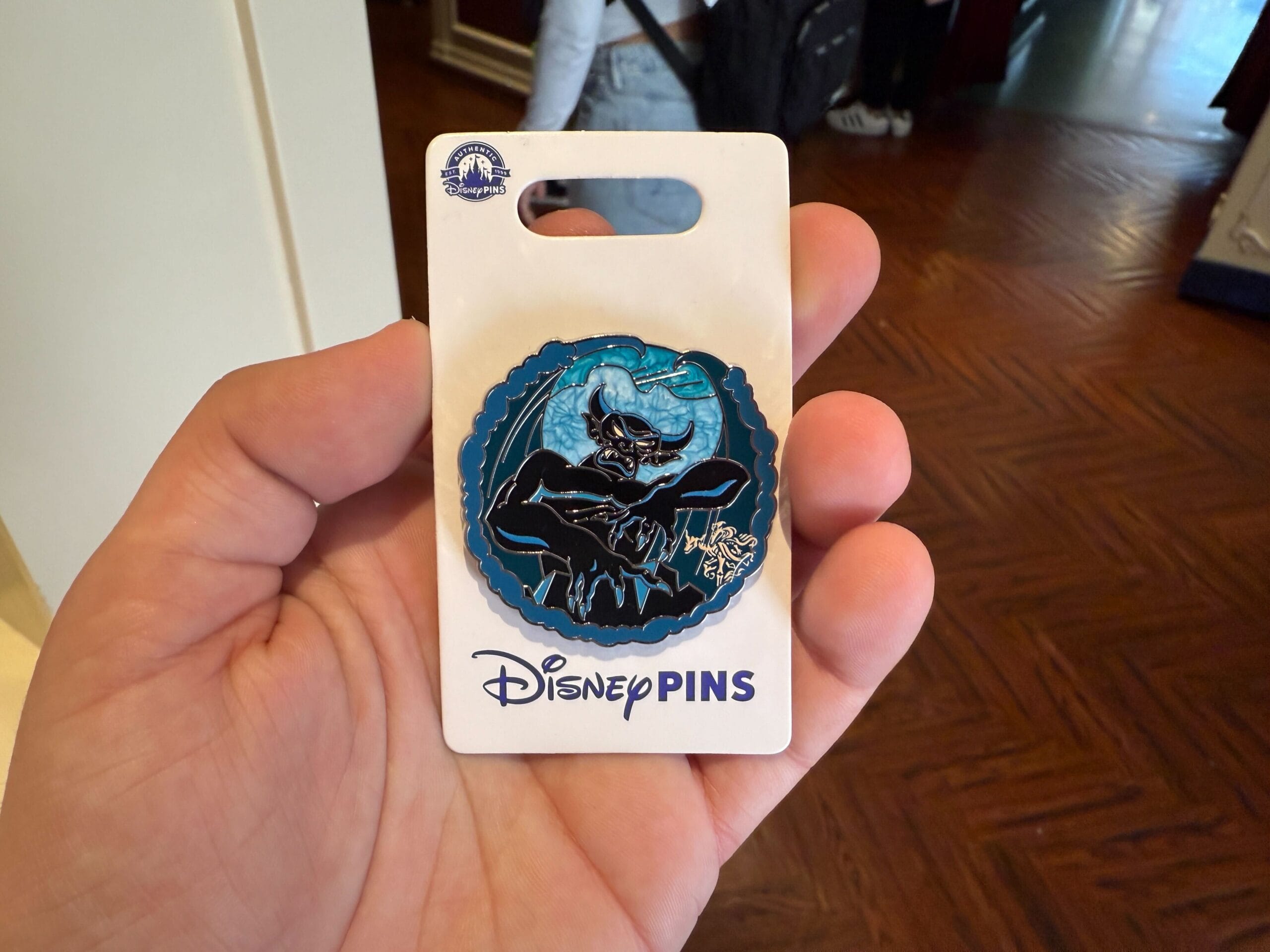 A hand holds a Disney pin featuring a winged creature in front of a blue background, attached to packaging labeled "Disney Pins.